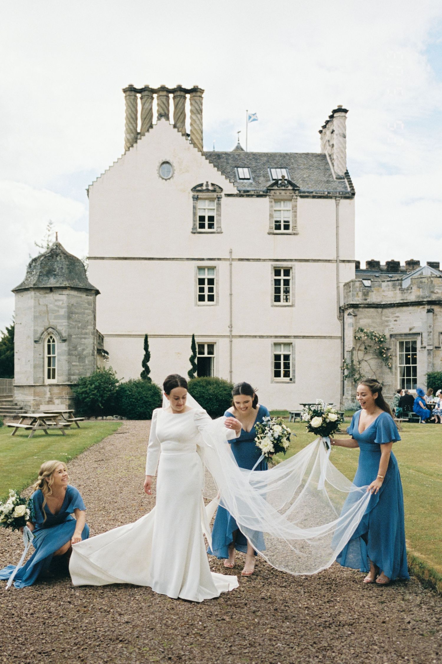 Bridesmaids in blue dresses help arrange a wedding gown's train on castle grounds in a series of candid photos.