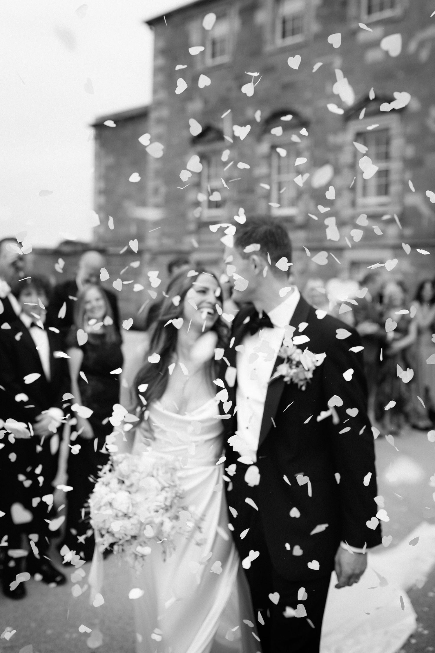 Black and white wedding photo showing couple walking through falling flower petals outside a brick building.