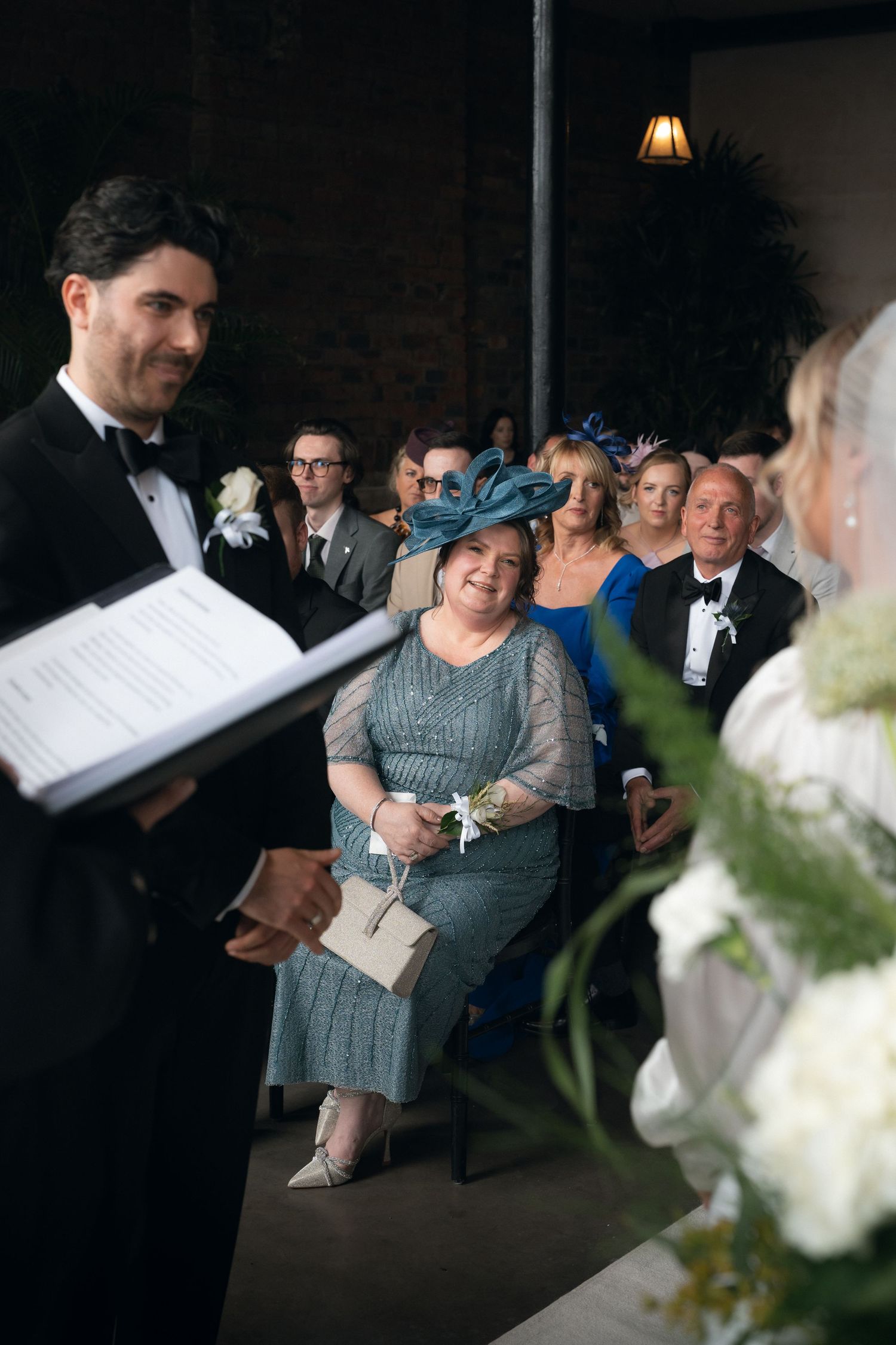 Wedding guests smile during ceremony, woman in blue hat and gray dress seated in foreground, officiant reads from book.