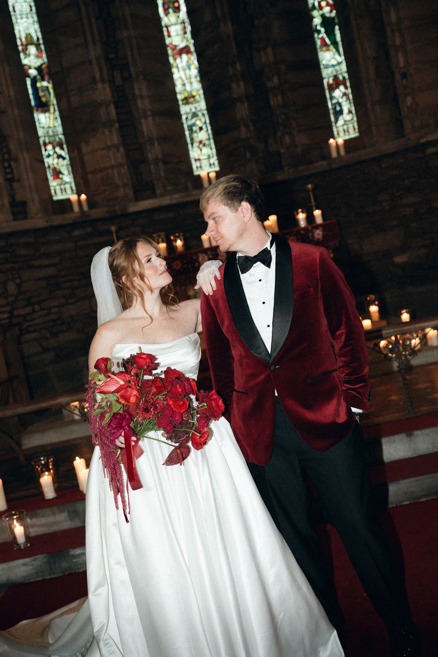 Newlyweds in a church with stained glass windows, bride in white gown and groom in burgundy velvet tuxedo with red bouquet.