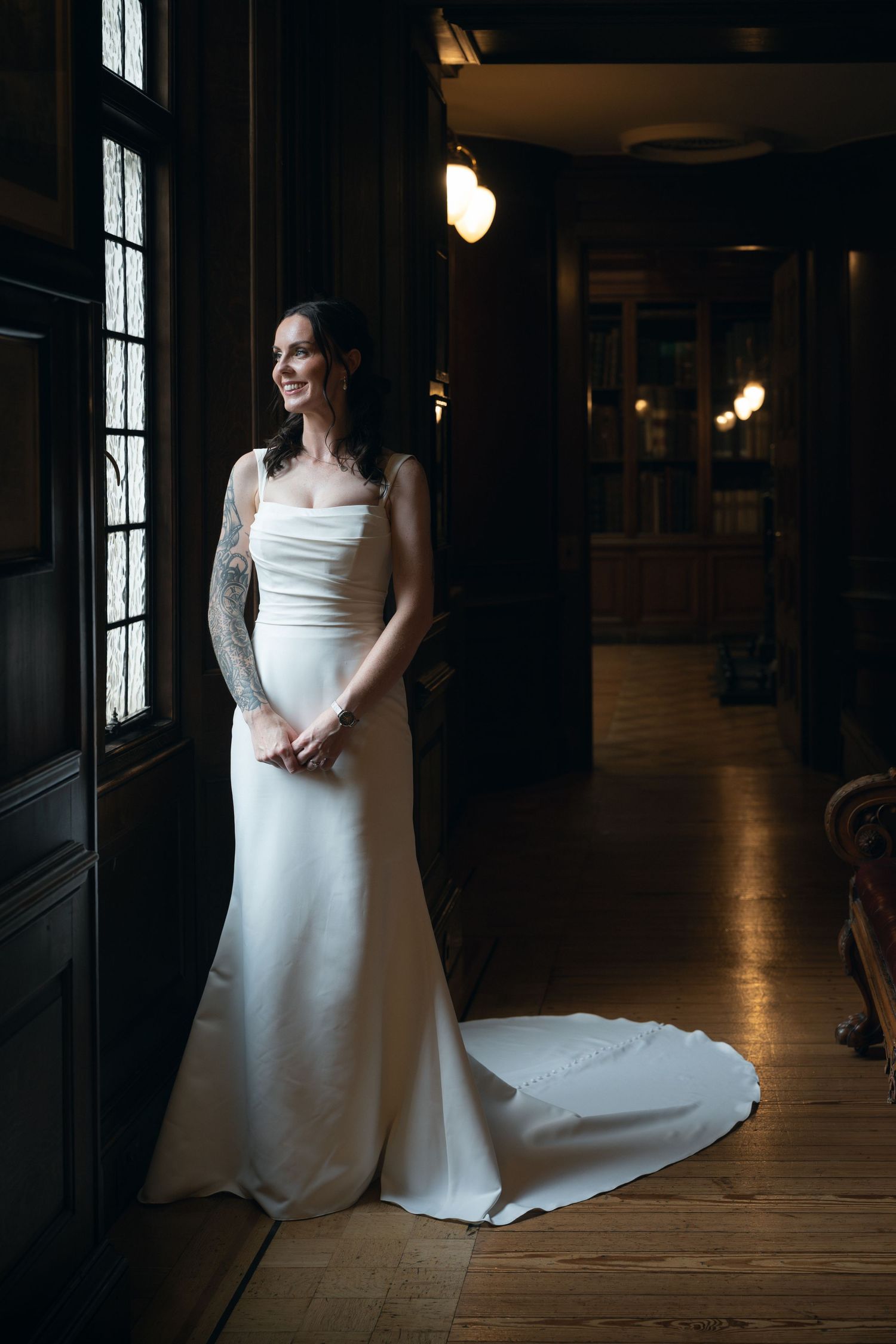 A figure in a flowing white wedding dress poses in a dark indoor space with dramatic window lighting.