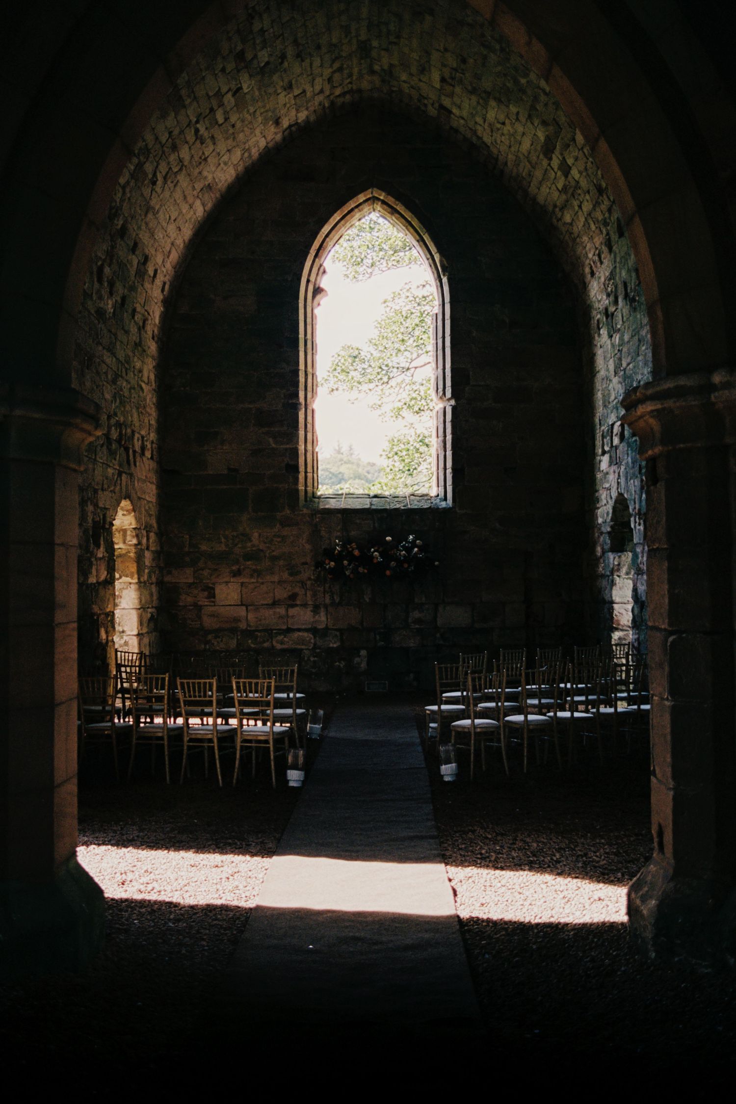 Light streams through arched windows in a dark stone church interior with rows of empty wooden chairs.