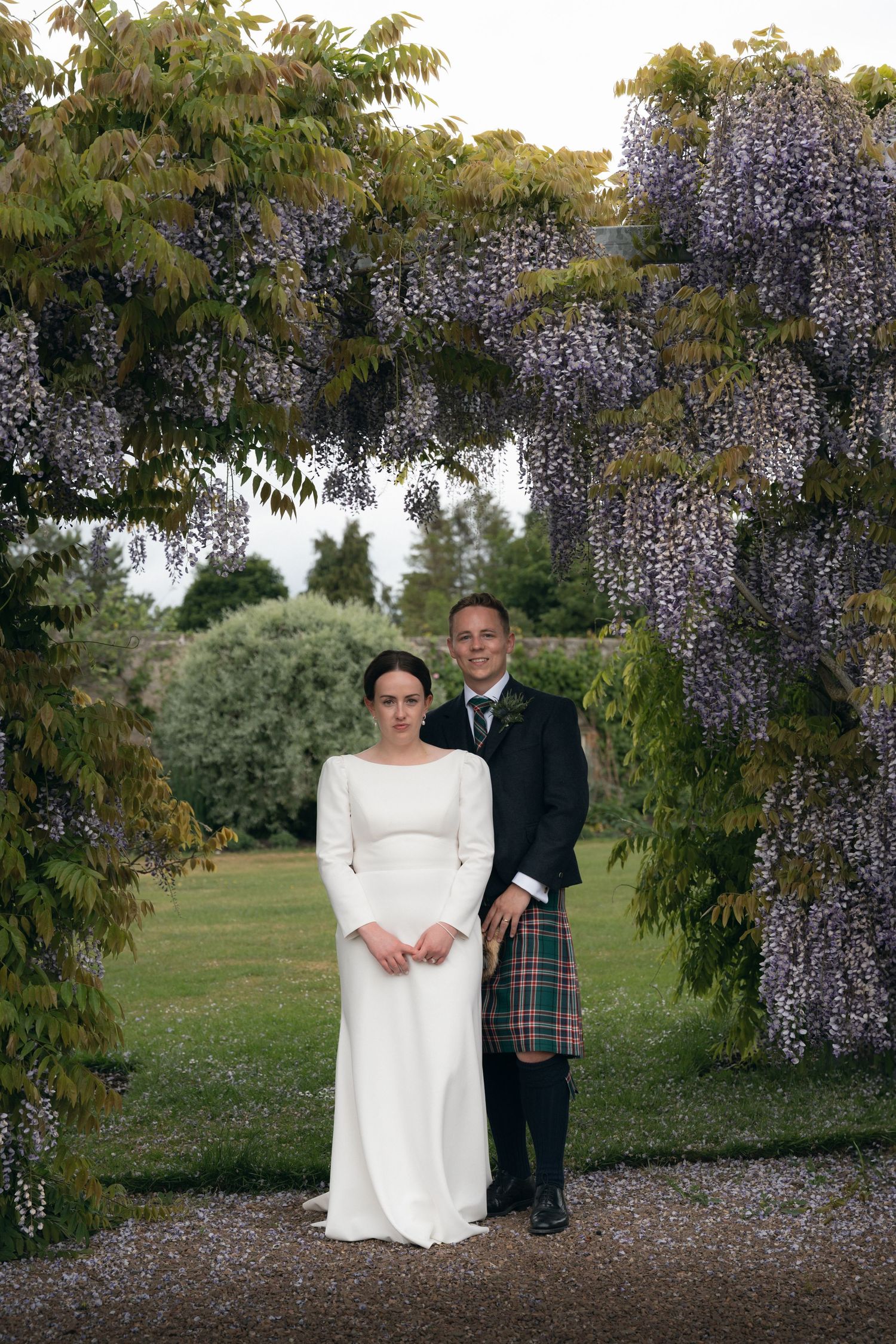 A couple poses under blooming wisteria vines in a garden setting during their wedding celebration.