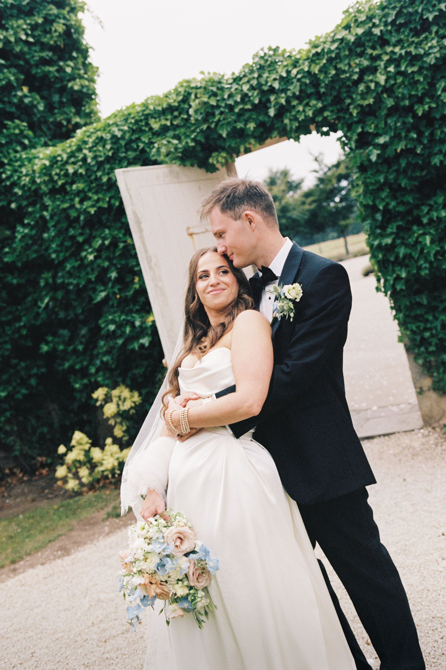 A romantic wedding couple embraces on a garden path surrounded by lush green foliage.
