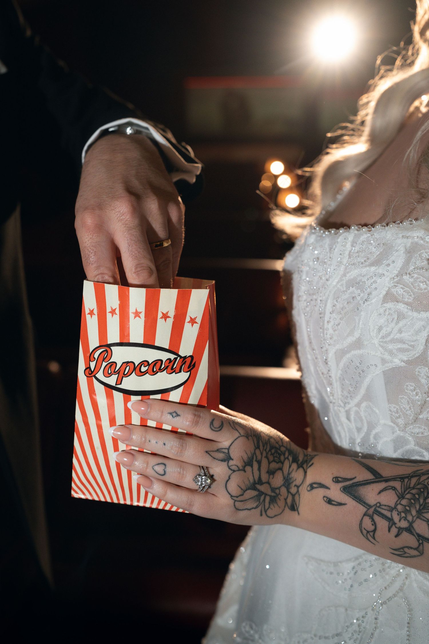 Bride and groom sharing a red and white striped popcorn box, bride showing floral tattoos and diamond ring.