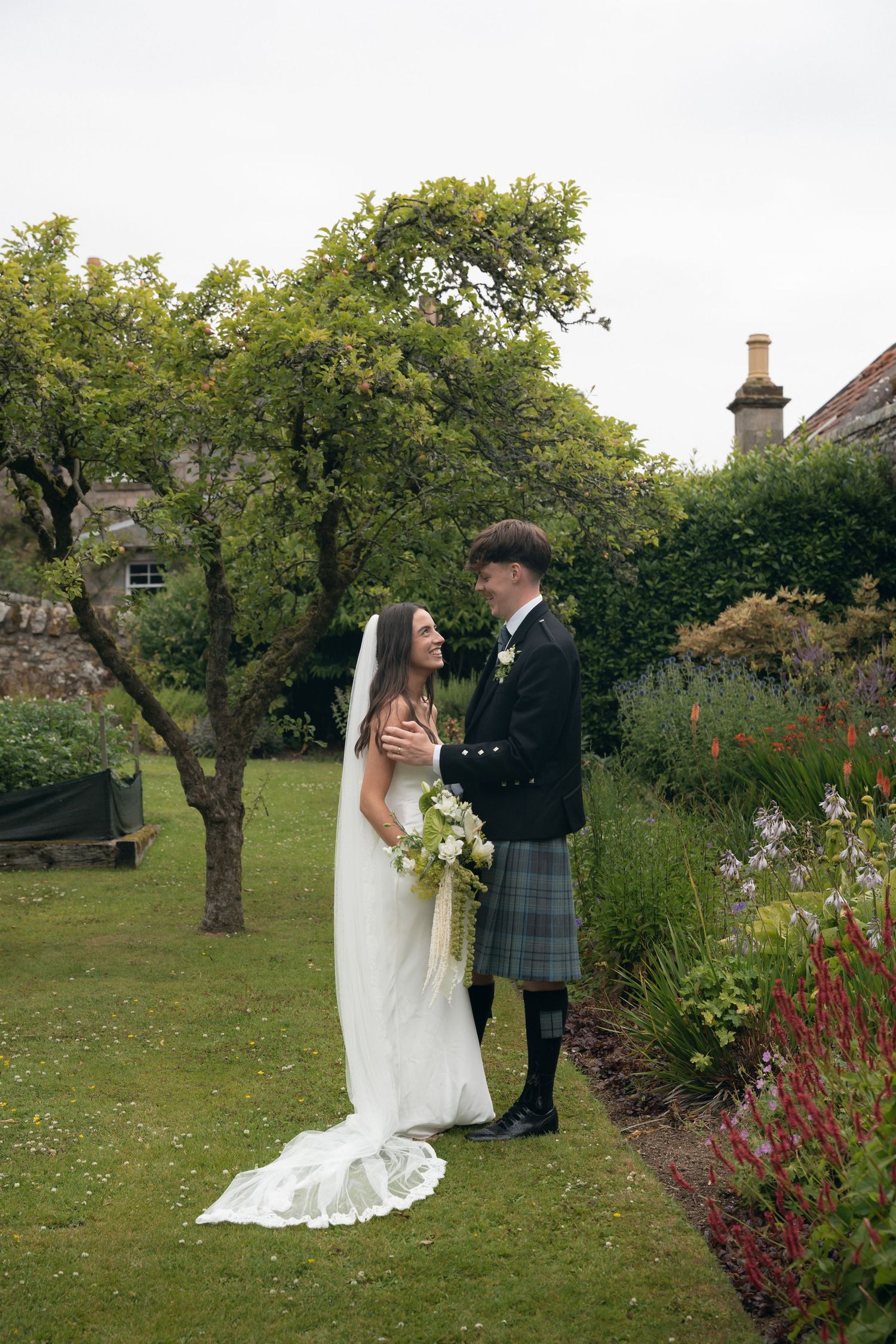 A couple in traditional Scottish wedding attire share romantic moments in a lush garden setting with blooming flowers.