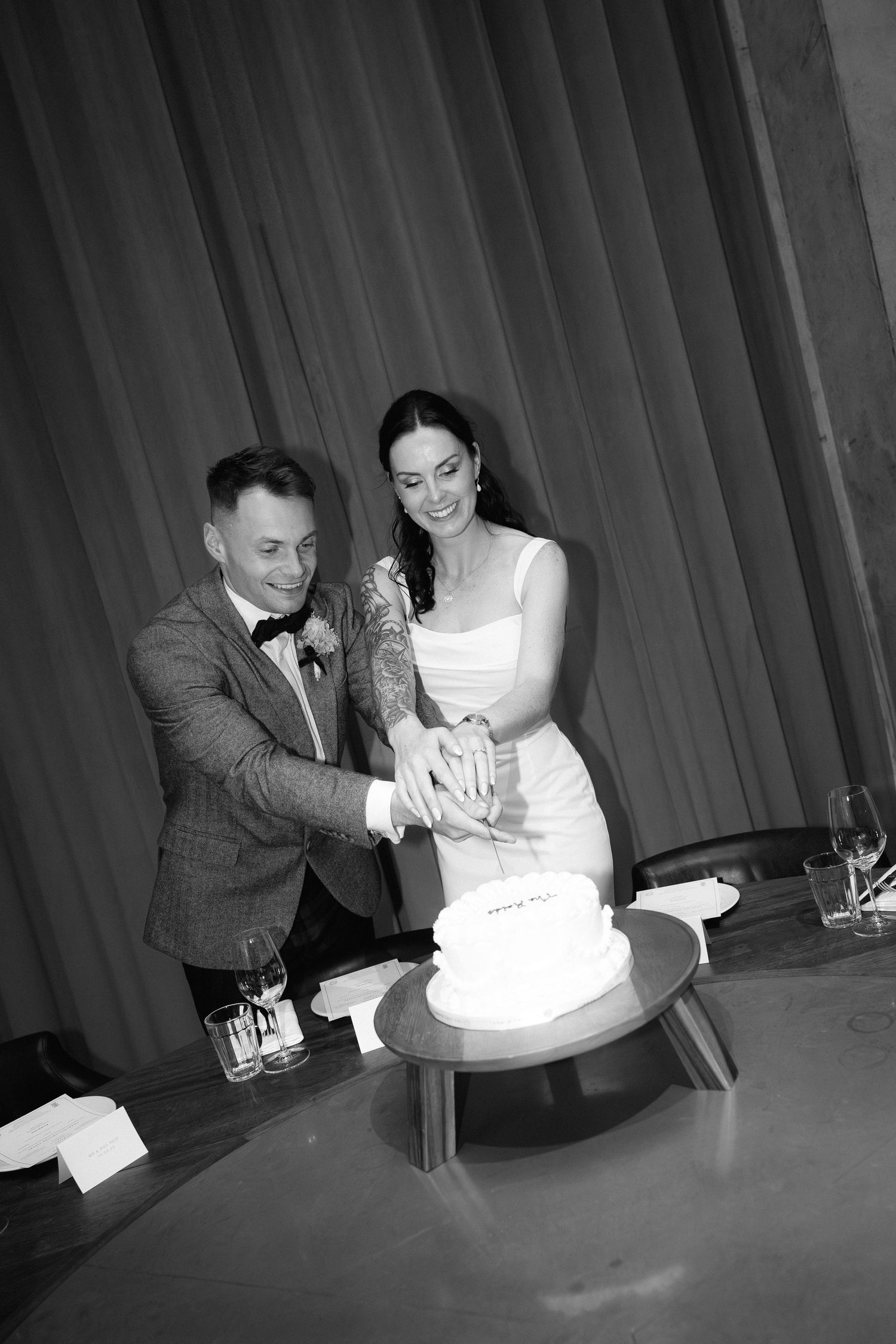 A black and white photo of newlyweds cutting their wedding cake together at their reception celebration.