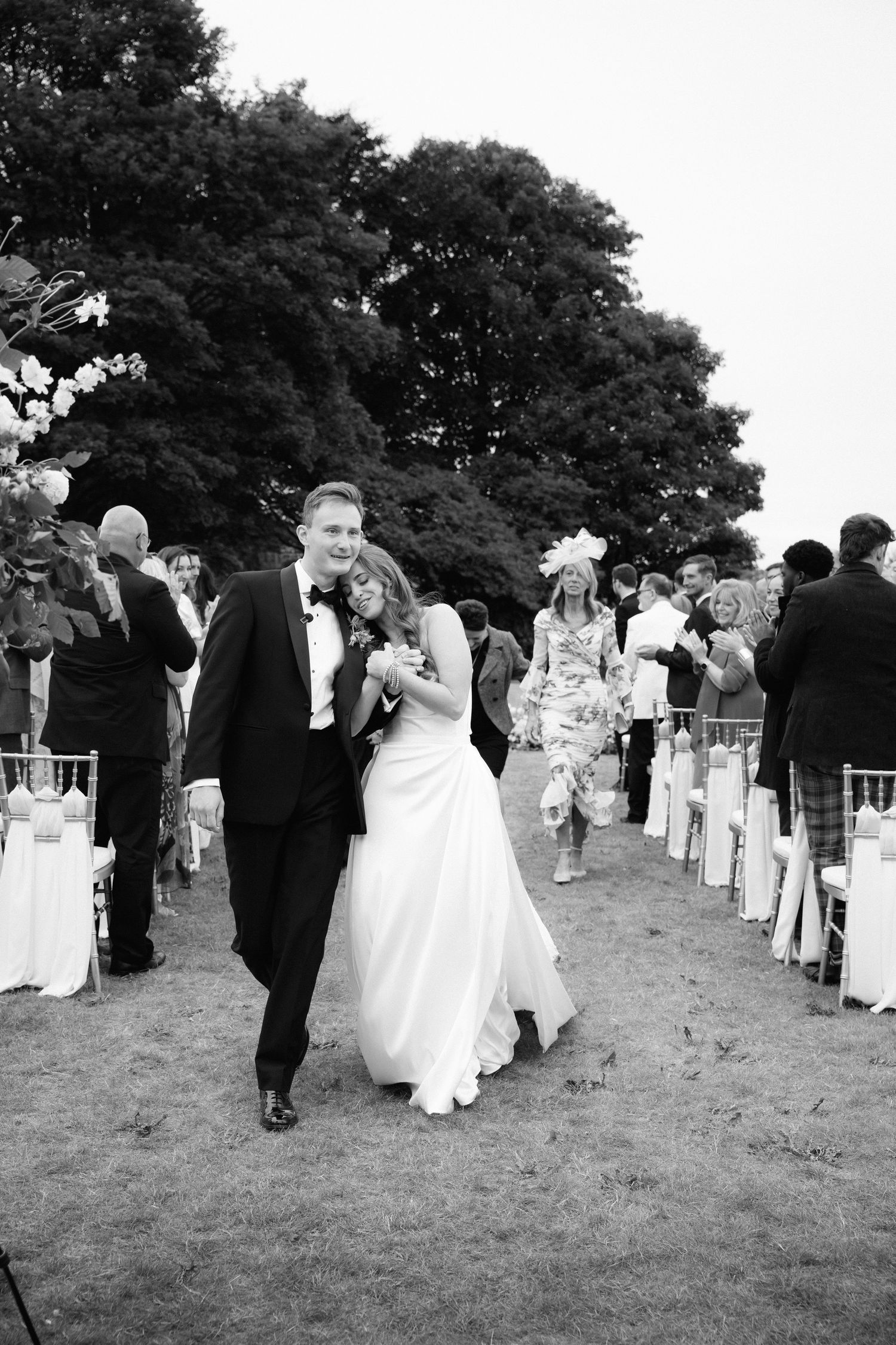 Black and white panoramic shot of wedding guests mingling on a grassy lawn during an outdoor reception.