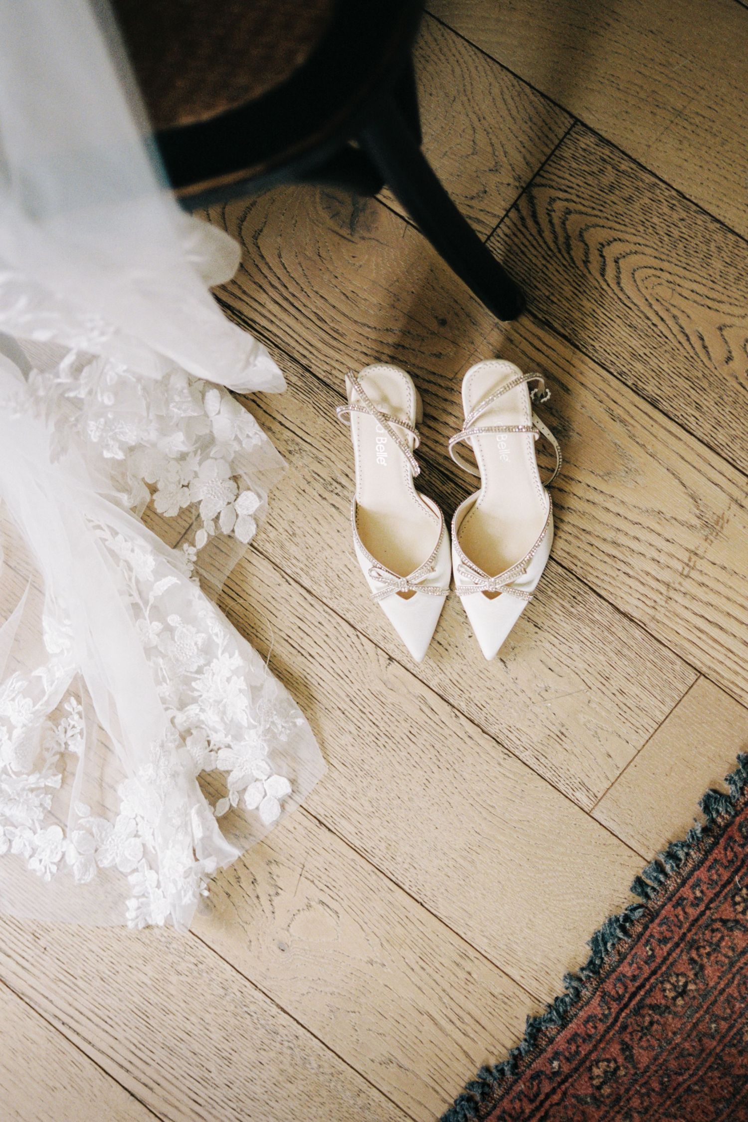 White bridal shoes and flowing veil fabric rest on a light hardwood floor next to an ornate patterned rug.