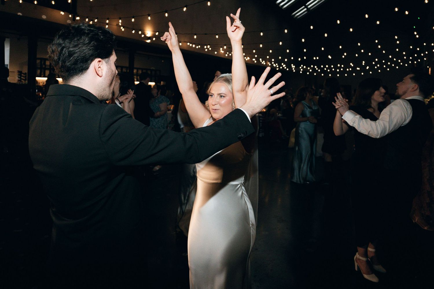 Couple dancing elegantly at a wedding reception under warm string lights in a dimly lit venue.
