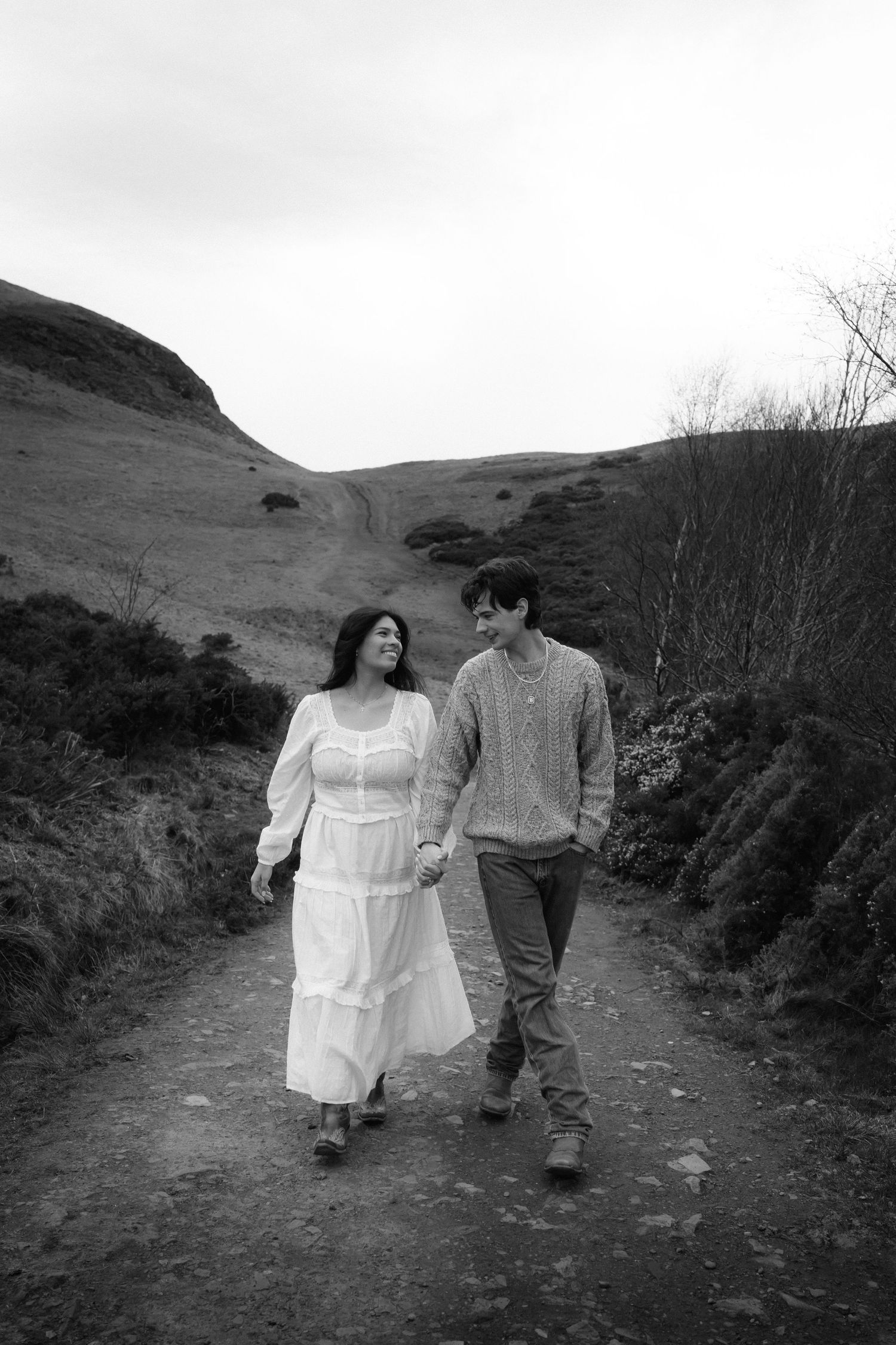 Black and white photo of a couple holding hands walking on a scenic mountain path, woman in white dress.