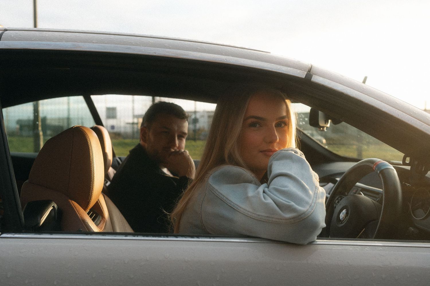 Two people sit in a car with tan leather interior during golden hour lighting.