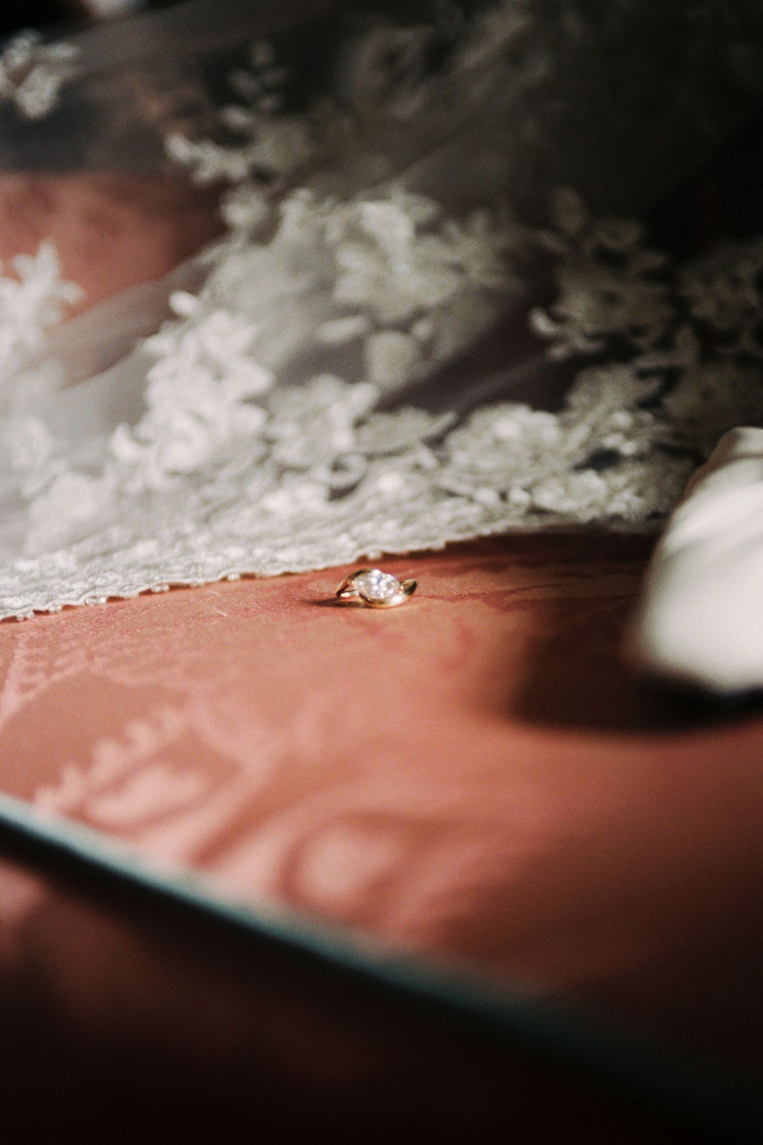 A delicate diamond engagement ring rests beside white lace on a red surface in soft lighting.