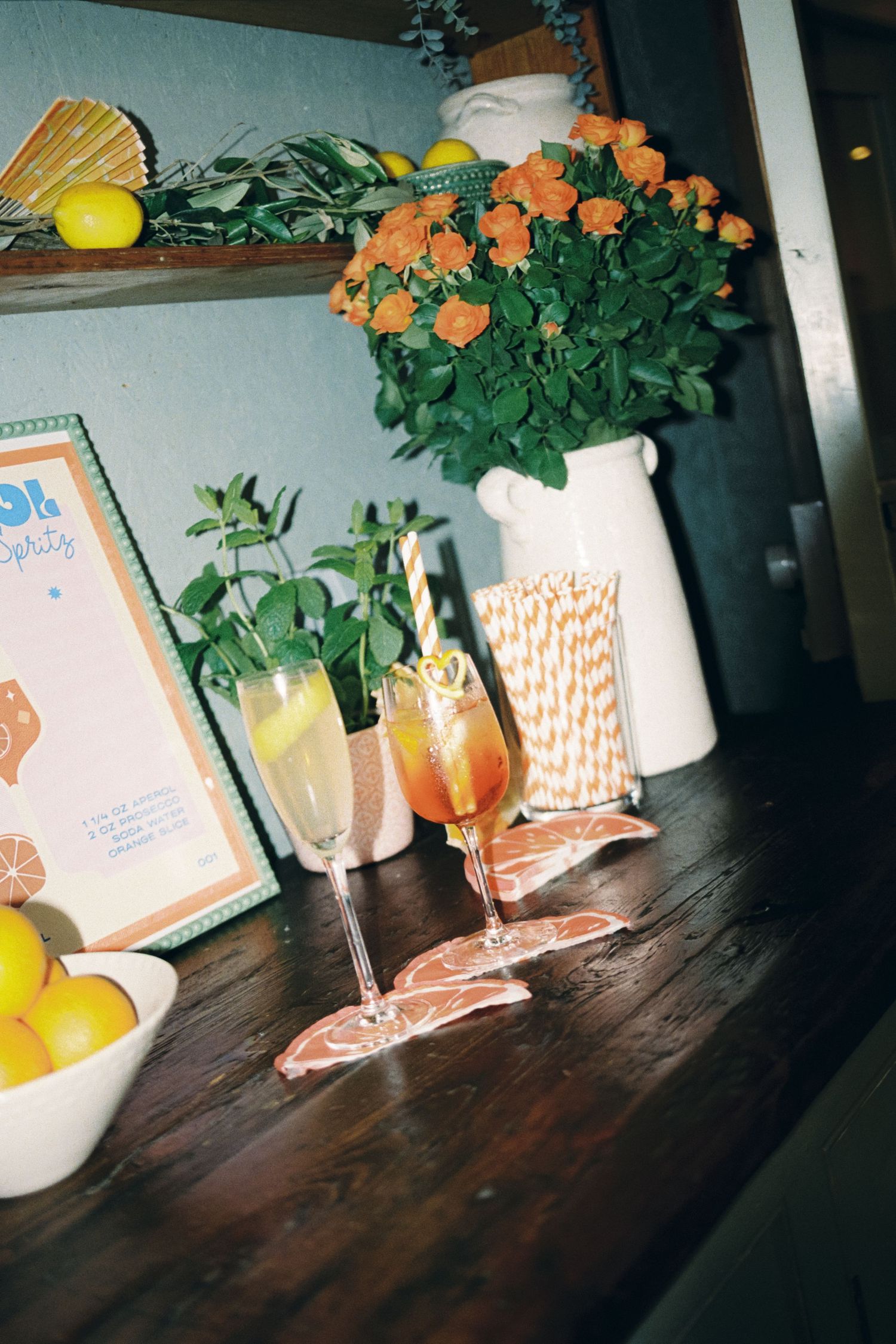 Two cocktail glasses on a wooden surface with orange drinks, paper straws and a white vase holding orange flowers in background.