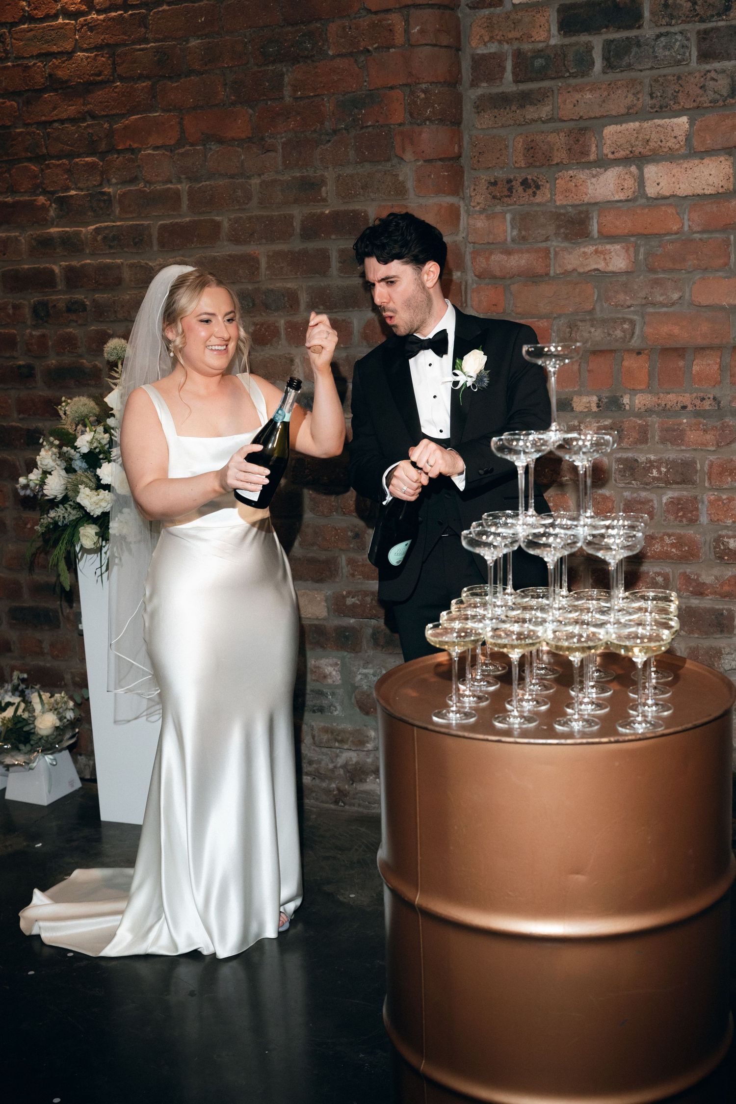 Bride and groom opening champagne beside a tiered glass tower at their wedding reception in a brick venue.