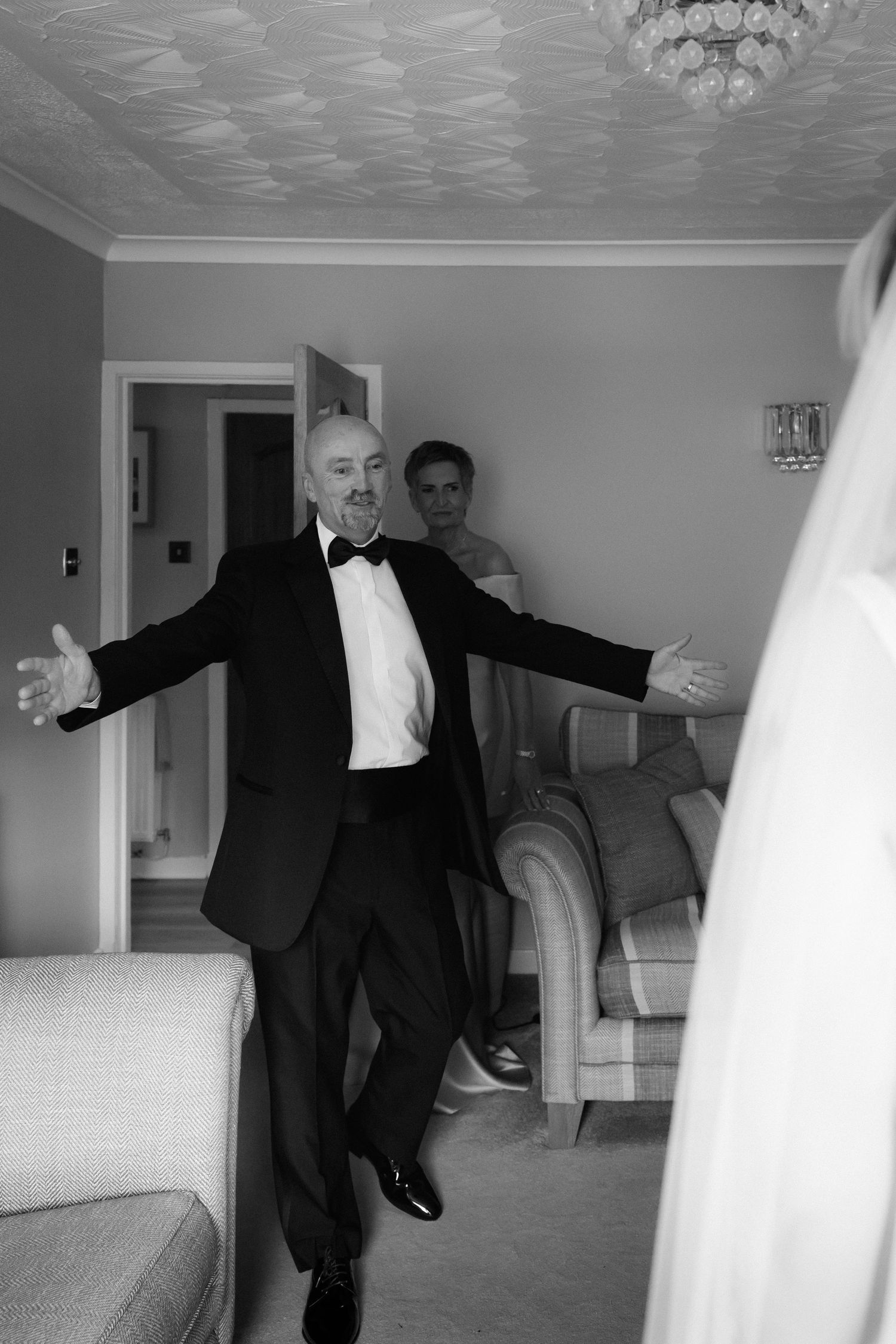 Black and white photo of a man in tuxedo with arms wide open, smiling joyfully inside a home on a wedding day.