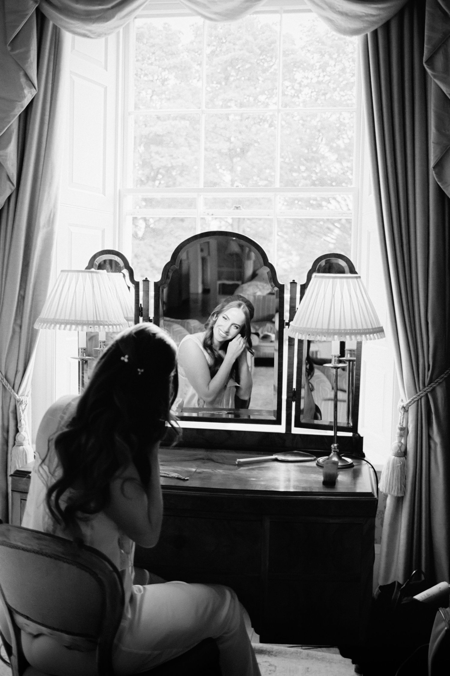 A vintage black and white photo showing a person seated at a desk near large windows with elegant curtains.