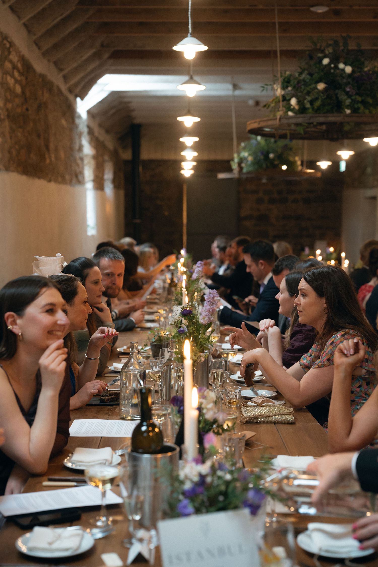 Guests enjoy a candlelit dinner at a long communal table decorated with floral centerpieces and wine glasses.