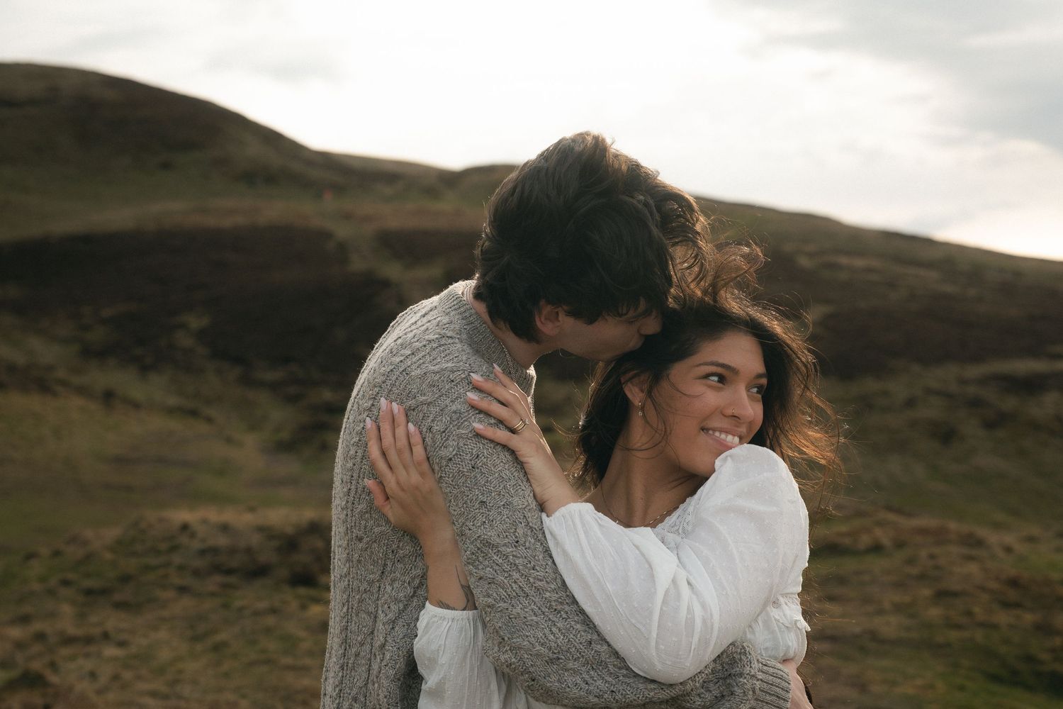 A couple embraces and smiles in a scenic green highland landscape under a cloudy sky.