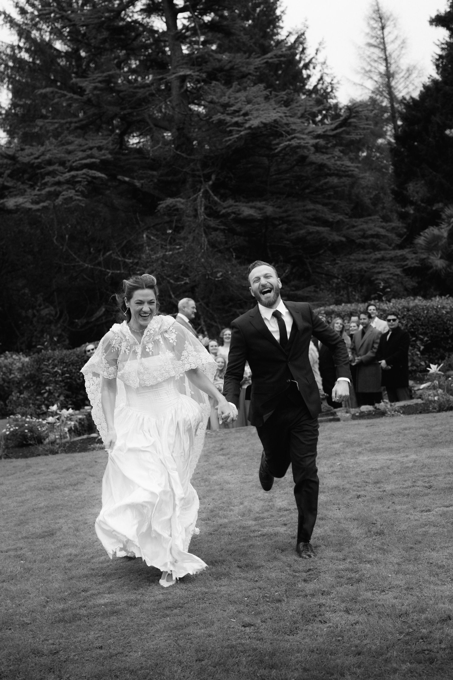 A bride in a flowing white dress runs playfully across a lawn with a suited companion at an outdoor wedding.