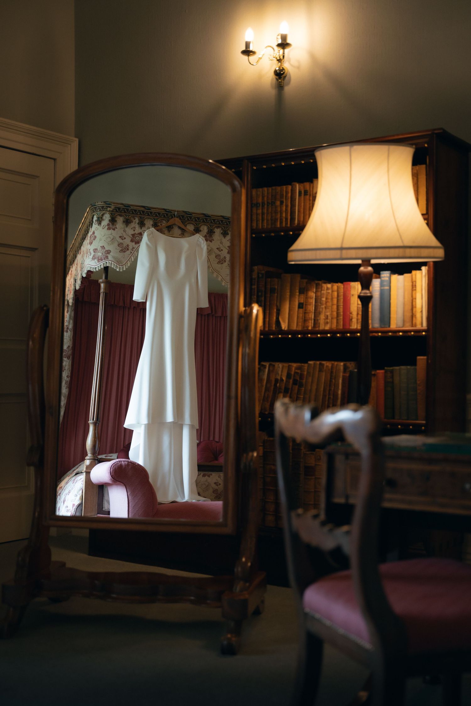 A white dress hangs illuminated in a dark room with antique furniture and bookshelves creating a moody atmosphere.