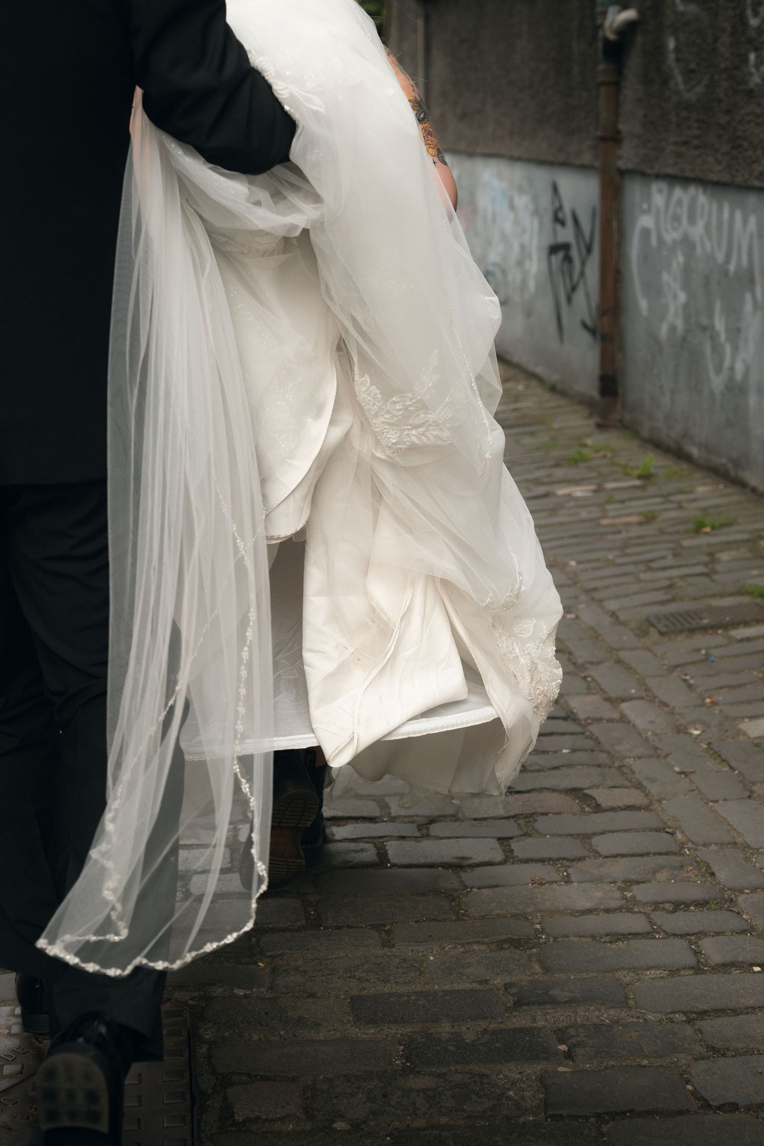 Groom carries bride's white wedding dress and veil along a cobblestone alley.