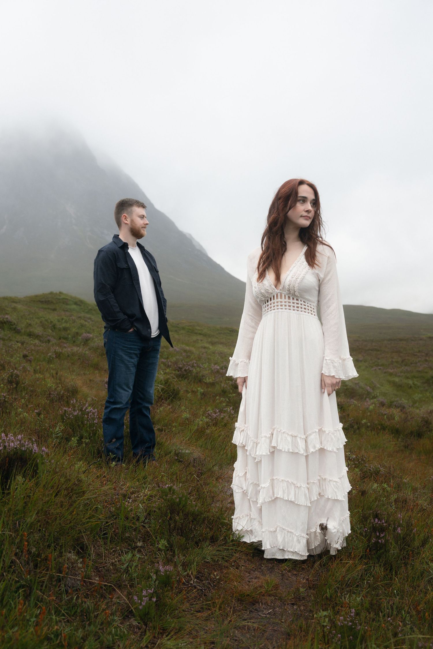A couple poses against a misty mountain backdrop in Scotland, with her in a bohemian white dress and him in dark attire.