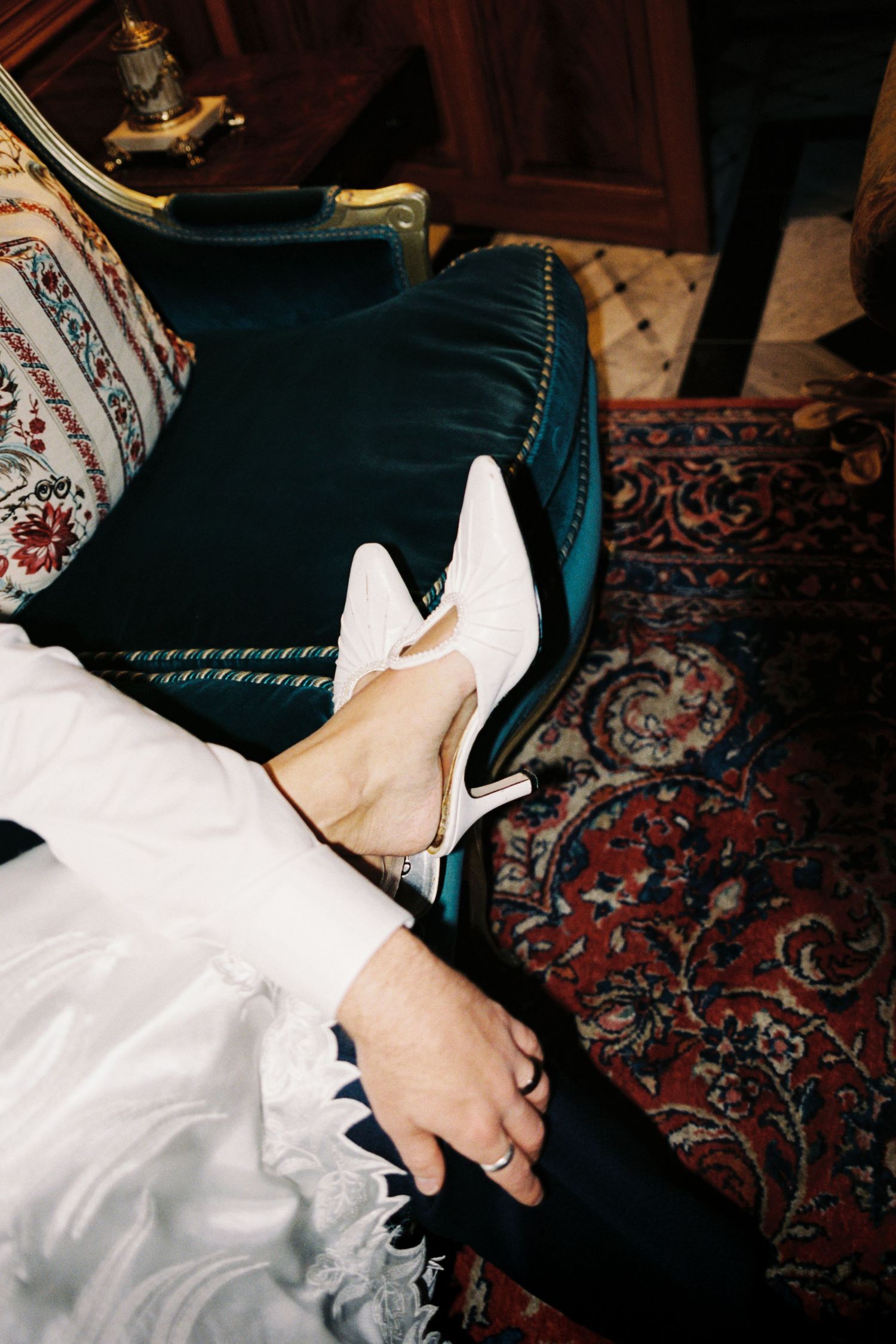 A person's hands adjust white high heel shoes while sitting on a patterned carpet.