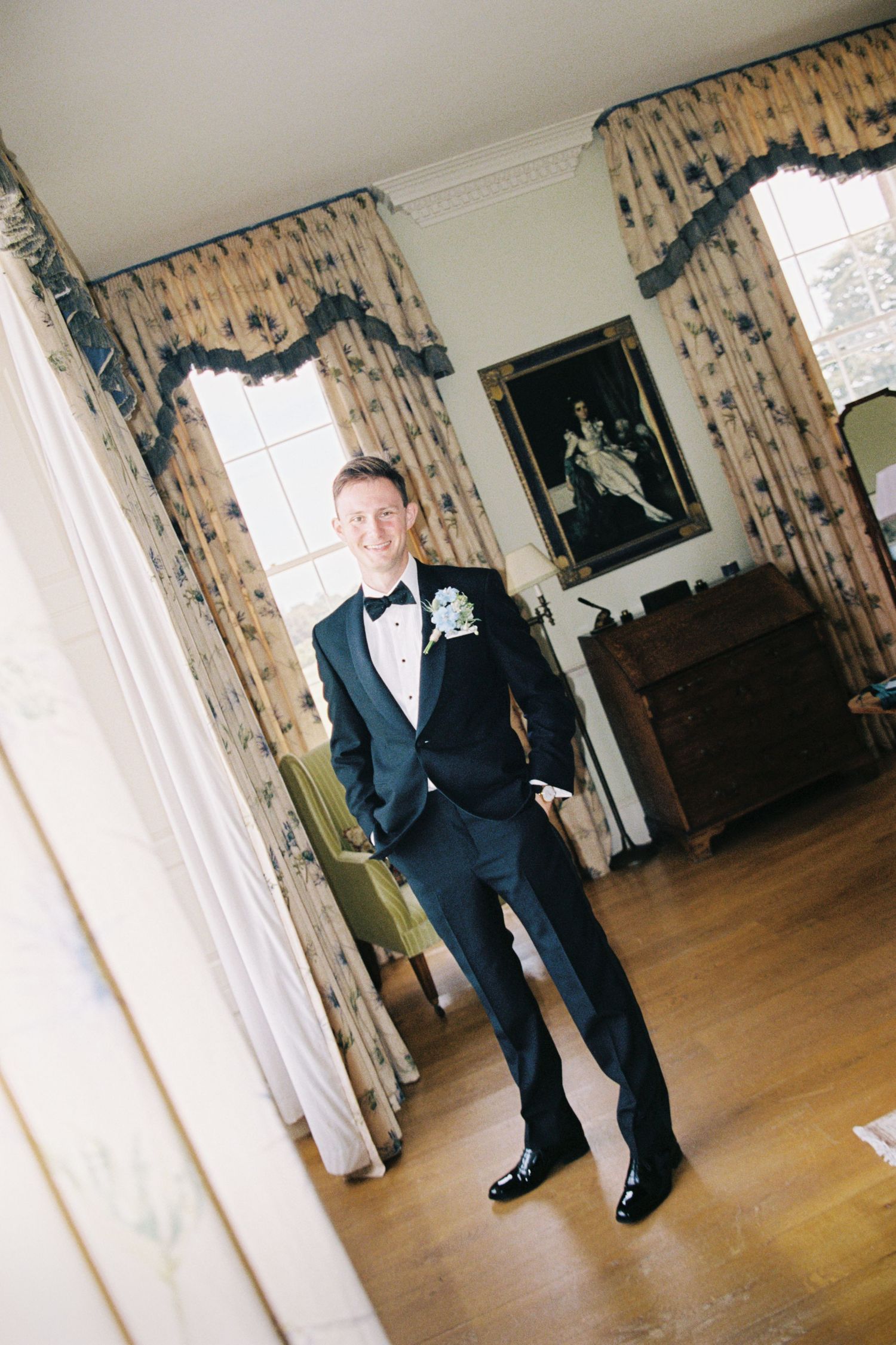 A person in a formal navy suit and bow tie stands on hardwood flooring near floral curtains in a bright room.