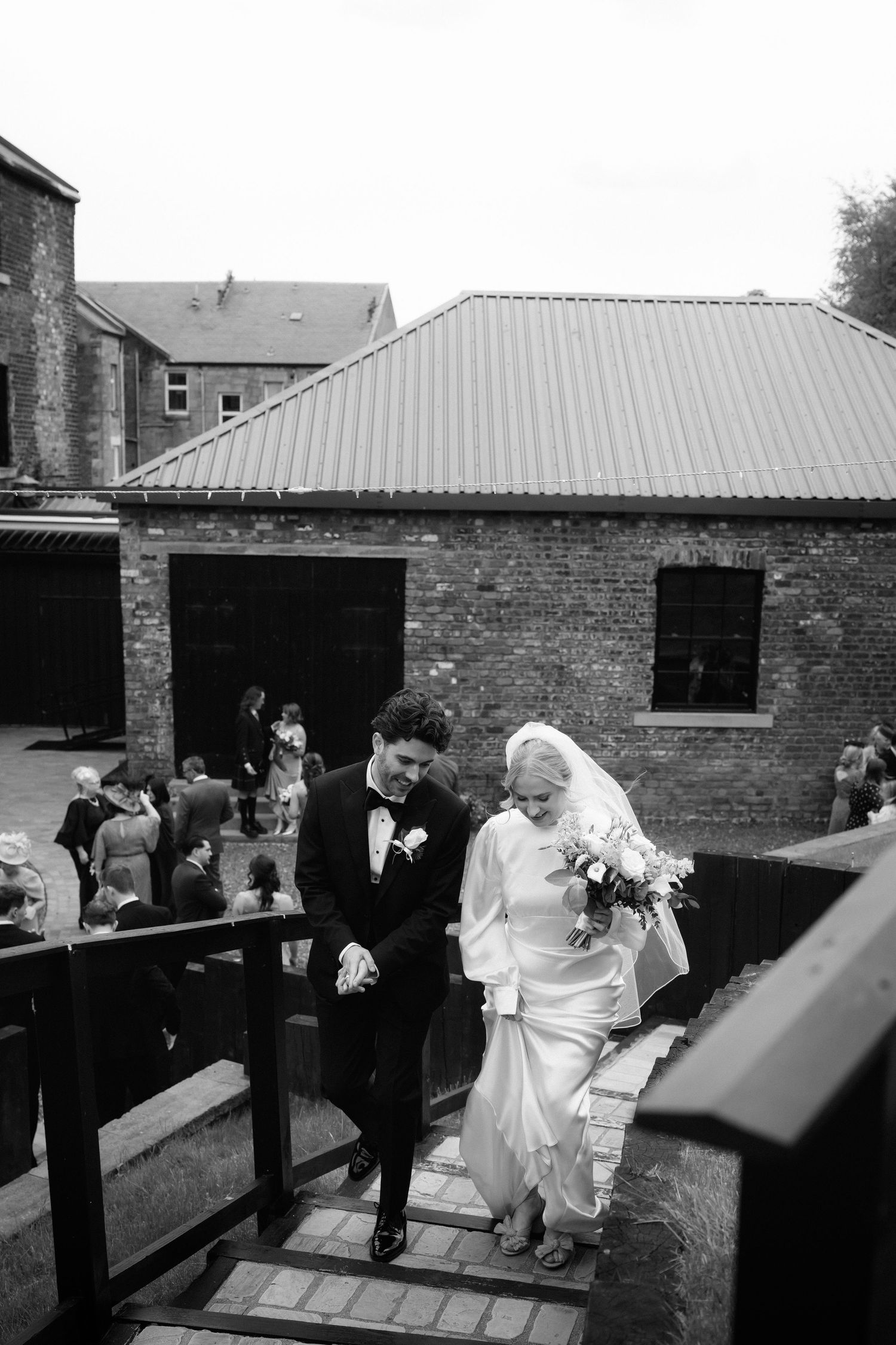 Black and white photo of a bride and groom descending outdoor steps at a brick venue, bride holding a bouquet.