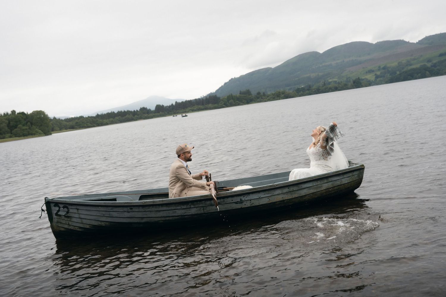 A romantic couple sits in a wooden boat on a serene lake surrounded by mountains on an overcast day.