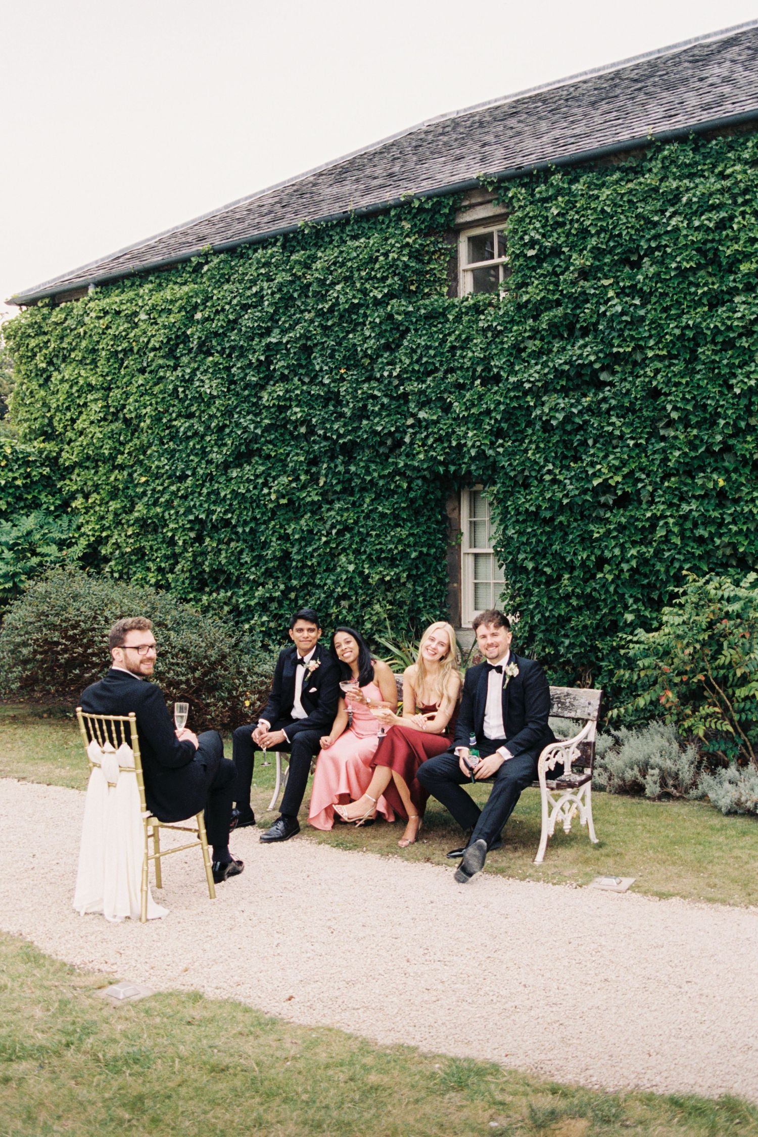 A group in formal attire sits together outside an ivy-covered building during a garden party event.
