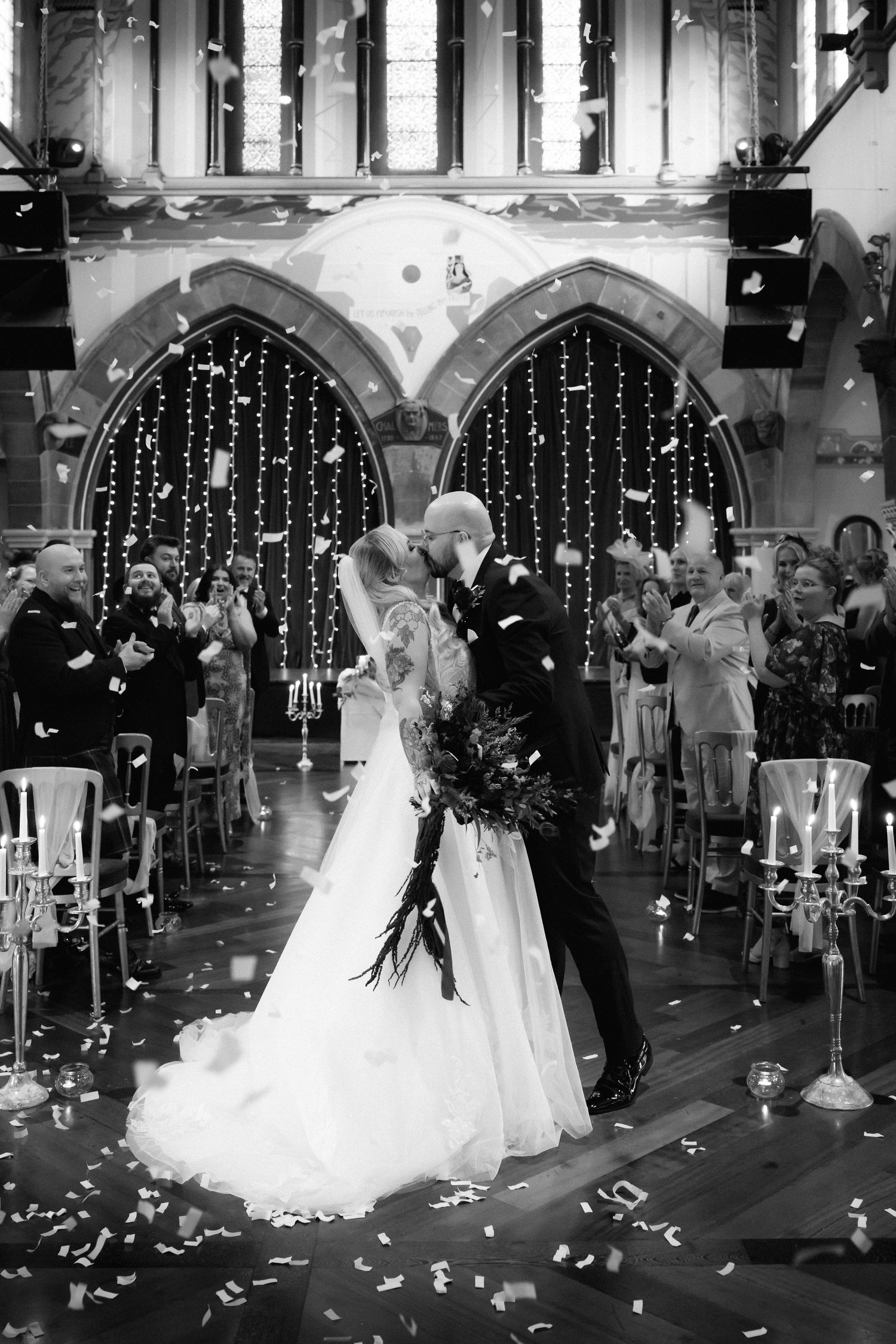 Black and white photo of bride and groom kissing at altar while guests throw confetti in elegant church.