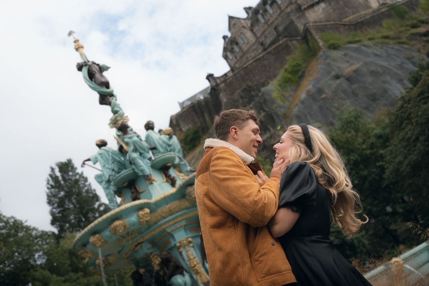 A couple embraces in a romantic moment against a scenic mountain backdrop on a cloudy day.