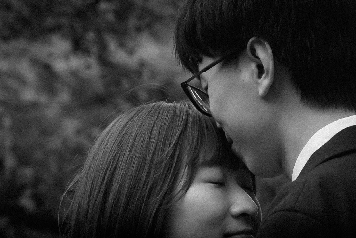 A black and white intimate romantic moment between a couple in glasses leaning close together outdoors.