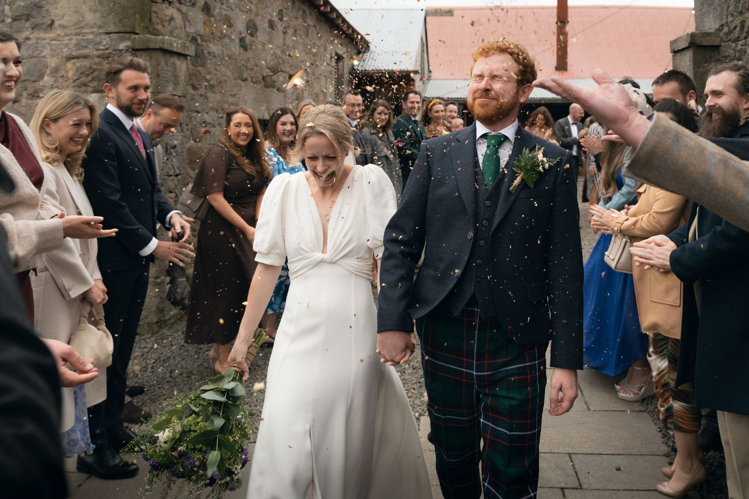 Wedding guests shower a joyful couple with confetti during an outdoor celebration at a stone venue.