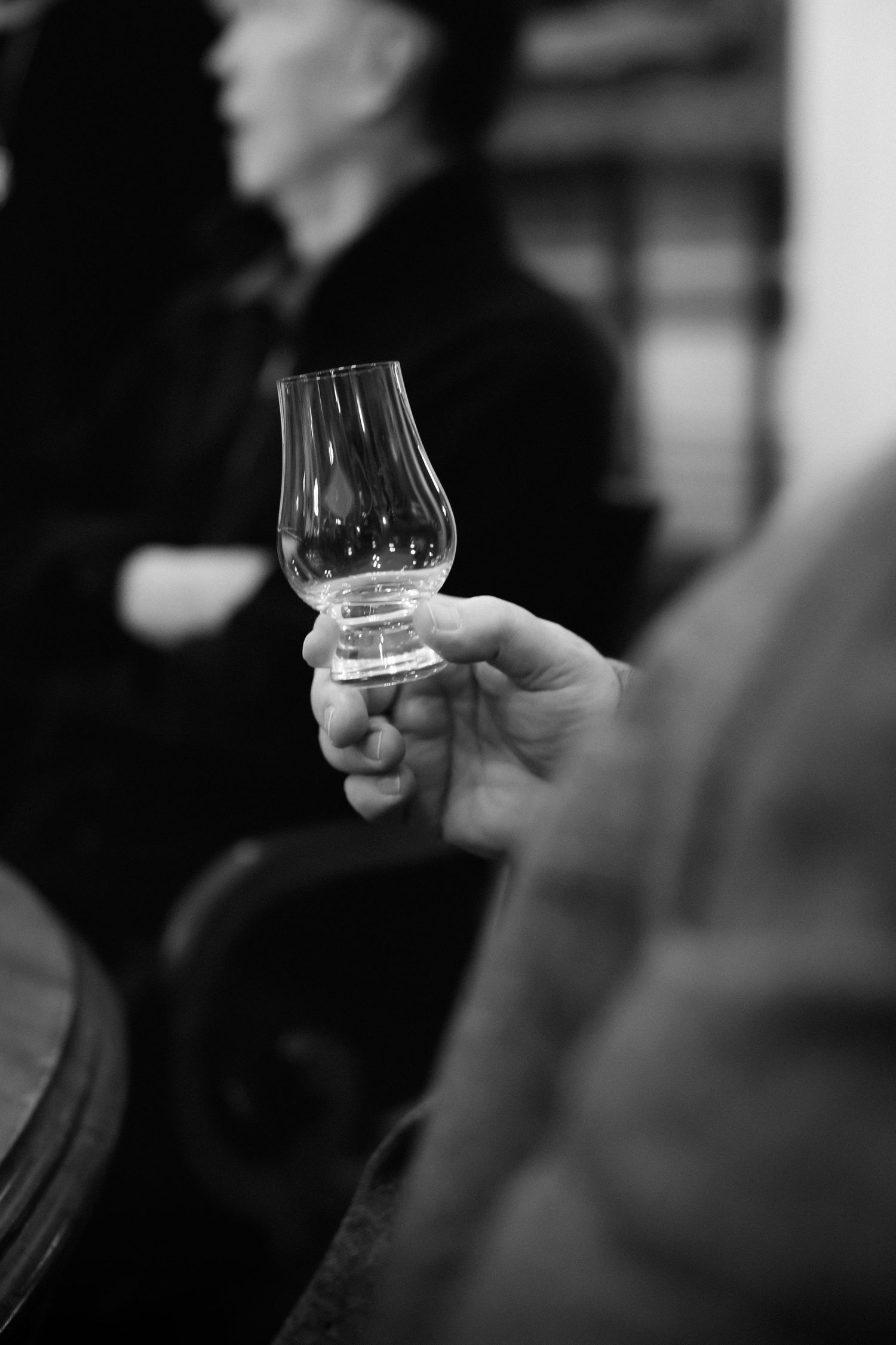 A hand holds a Glencairn whisky glass in black and white, with a blurred figure in the background.