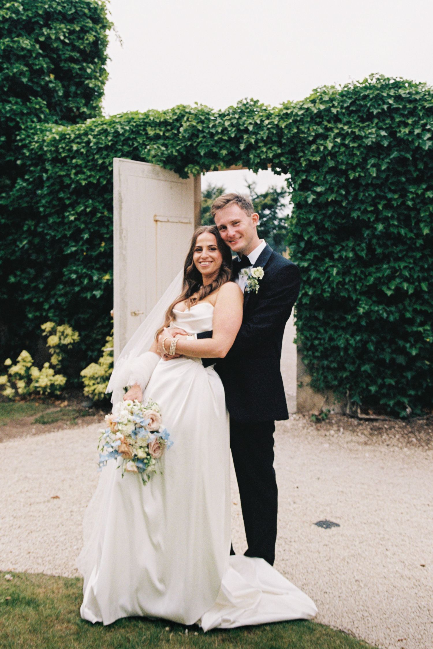 A couple in wedding attire poses in front of a white door surrounded by lush green ivy at an outdoor venue.