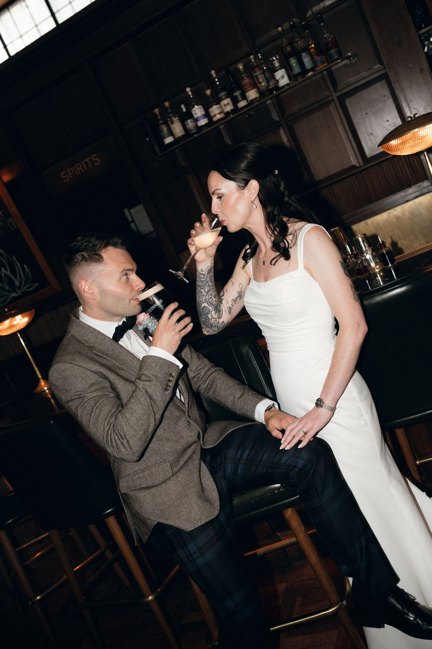 A couple in wedding attire share drinks at a bar counter with bottles displayed on shelves in the background.