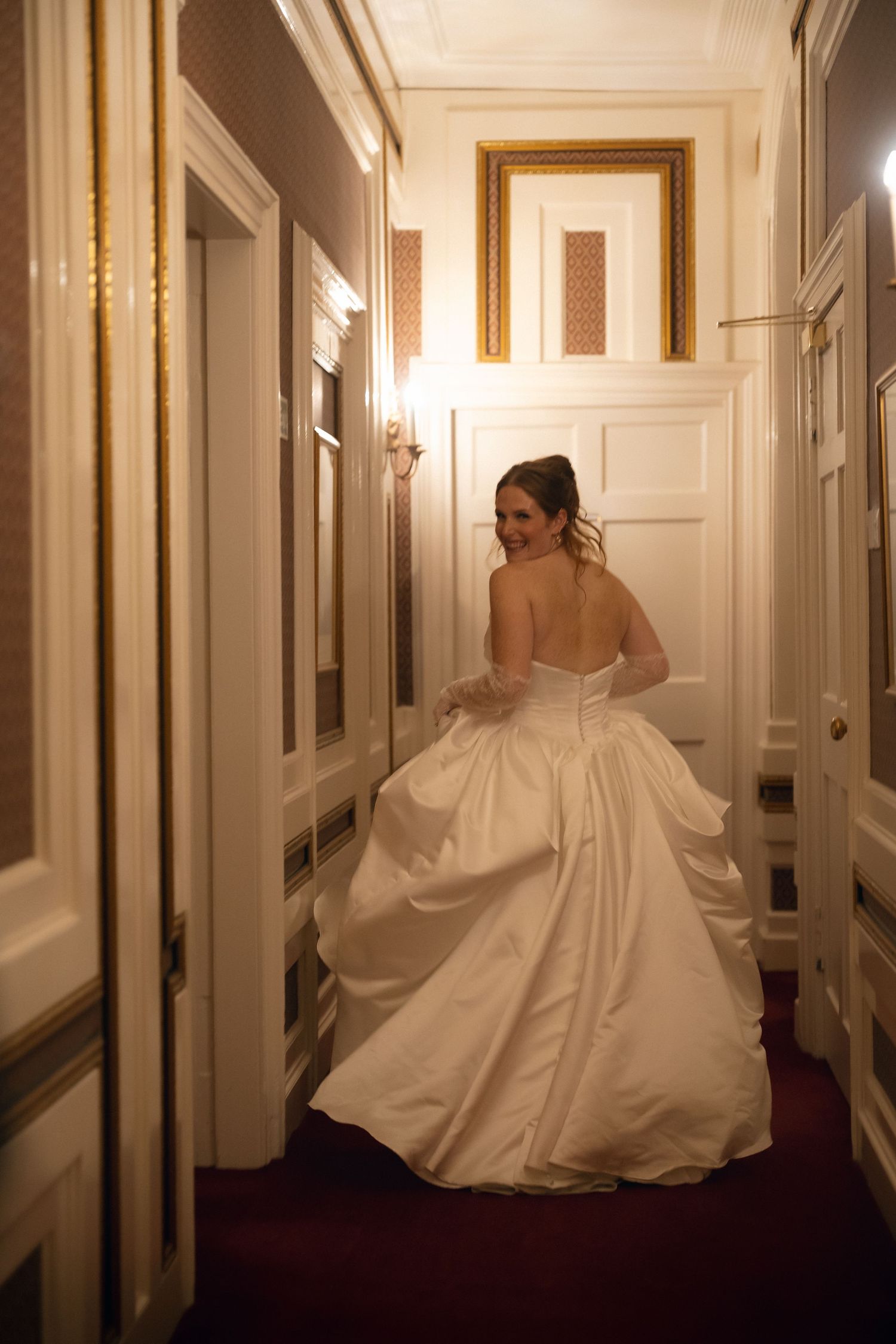 A bride in a flowing white ball gown walks down an elegant hallway with cream walls and burgundy carpet.