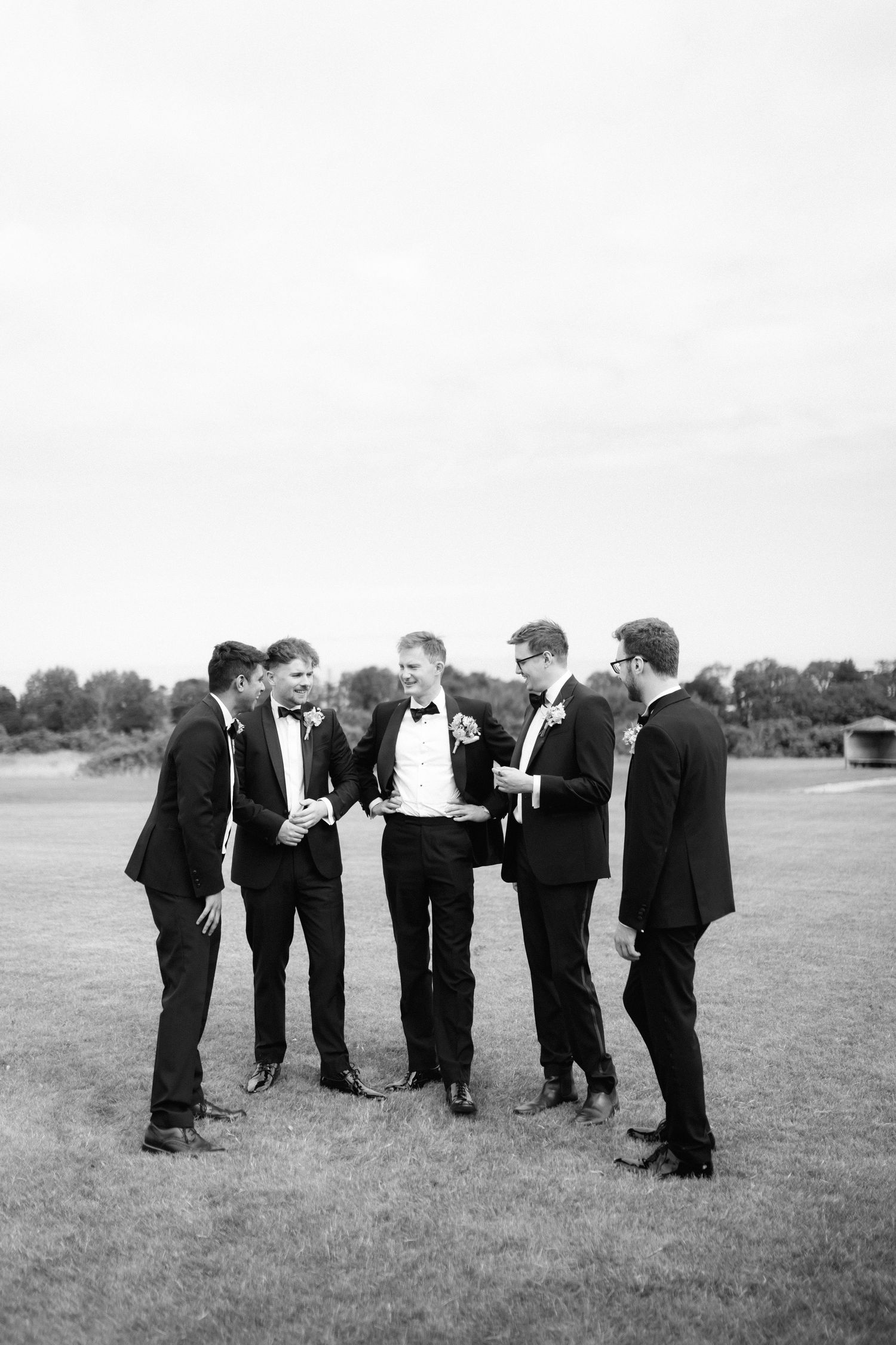 A group of groomsmen in black suits huddled together in a casual pose on a grassy field.