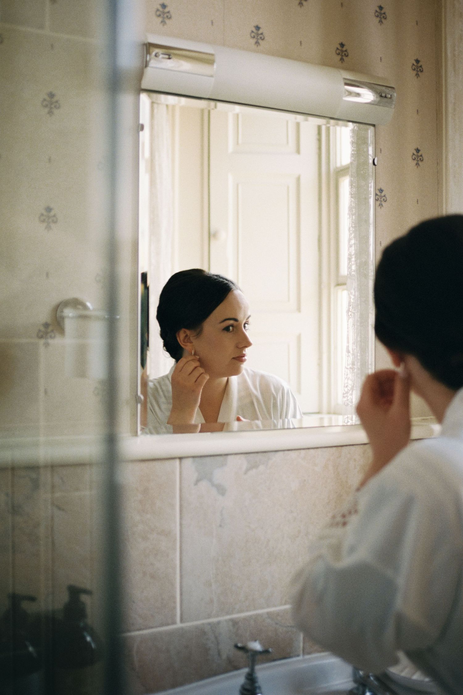 A reflection in a bathroom mirror shows someone gazing thoughtfully while getting ready in a vintage-style bathroom.