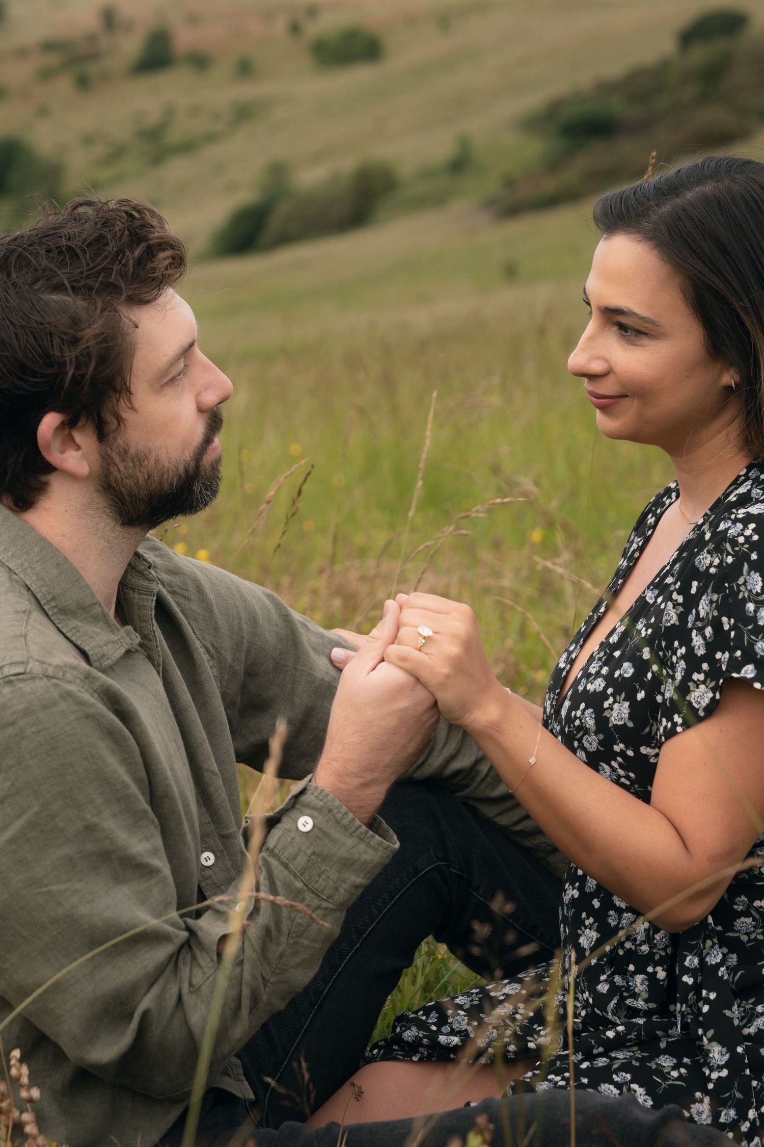 A couple holds hands while sitting in a grassy hillside meadow, sharing an intimate moment together.