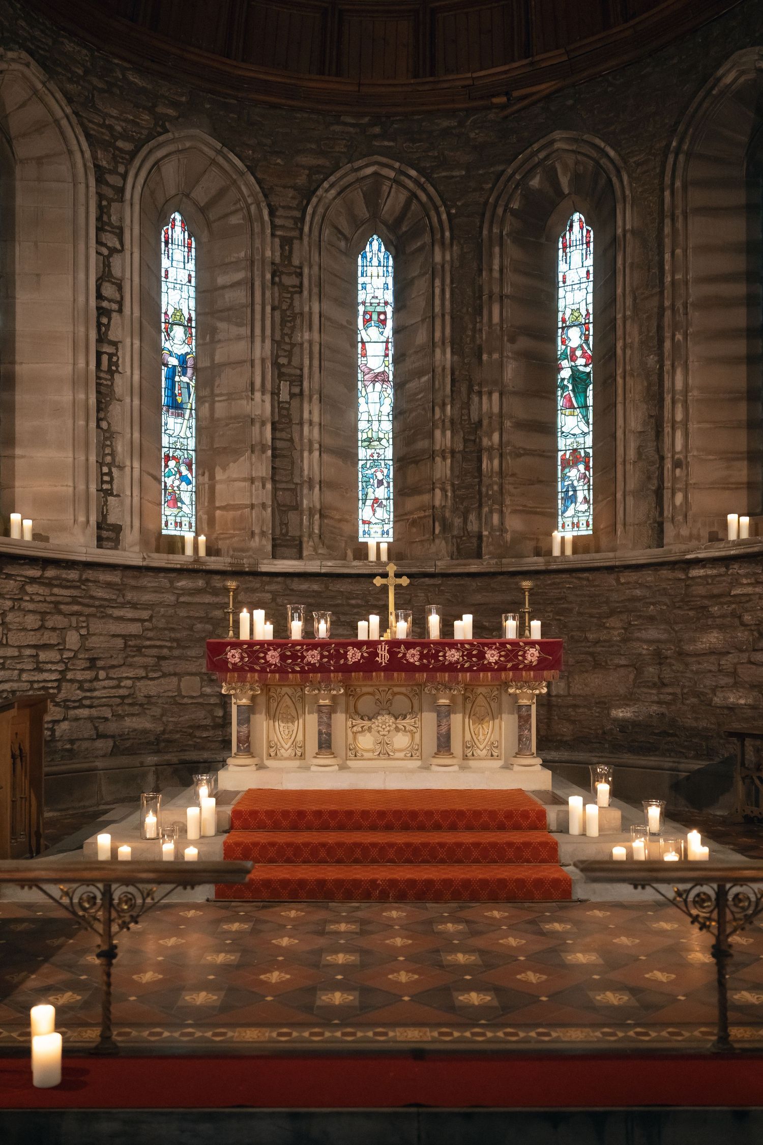 Stone church altar with stained glass windows, candles, and red carpeted steps leading to an ornate communion table.