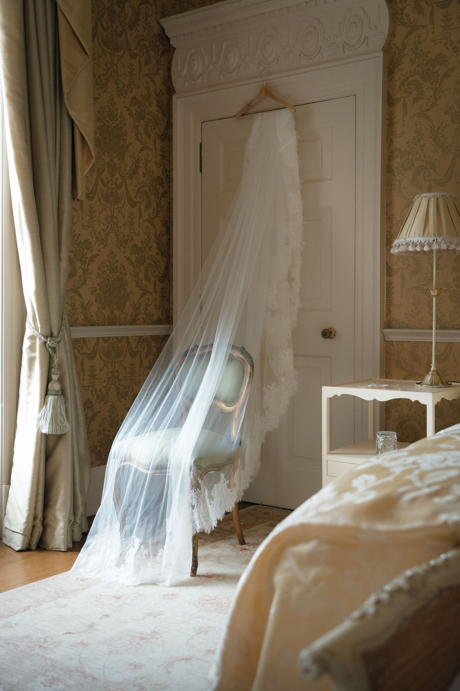 White wedding dress hanging elegantly in a vintage room with ornate wallpaper and soft natural lighting.