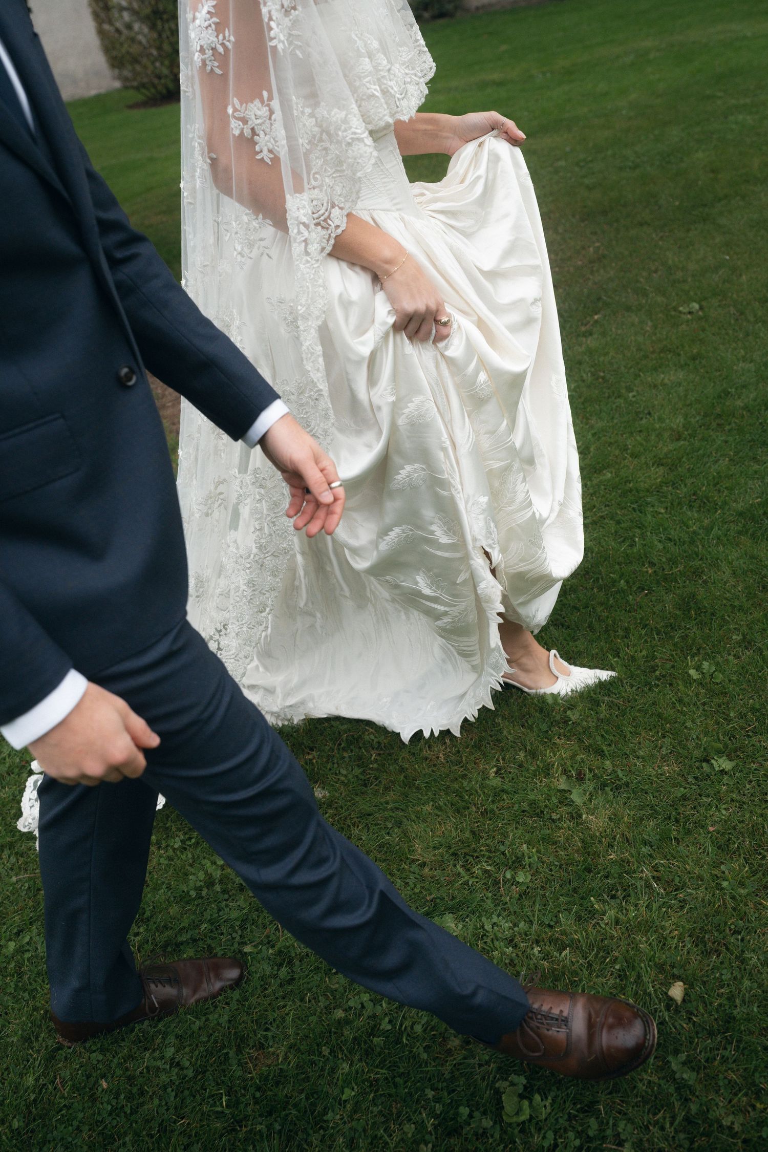 Wedding couple's feet play a playful game of footsie on green grass, showing dress hem and suit pants.