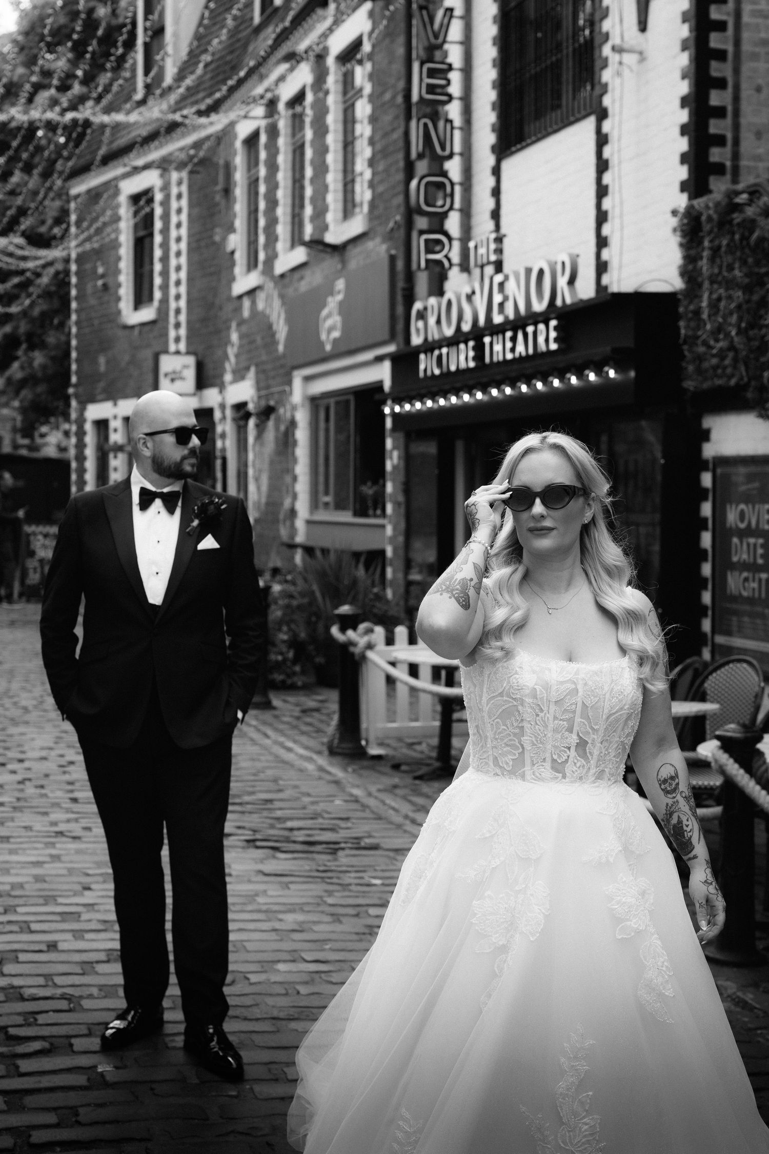 Bride adjusting sunglasses outside Grosvenor Picture Theatre with groom in black and white photo.