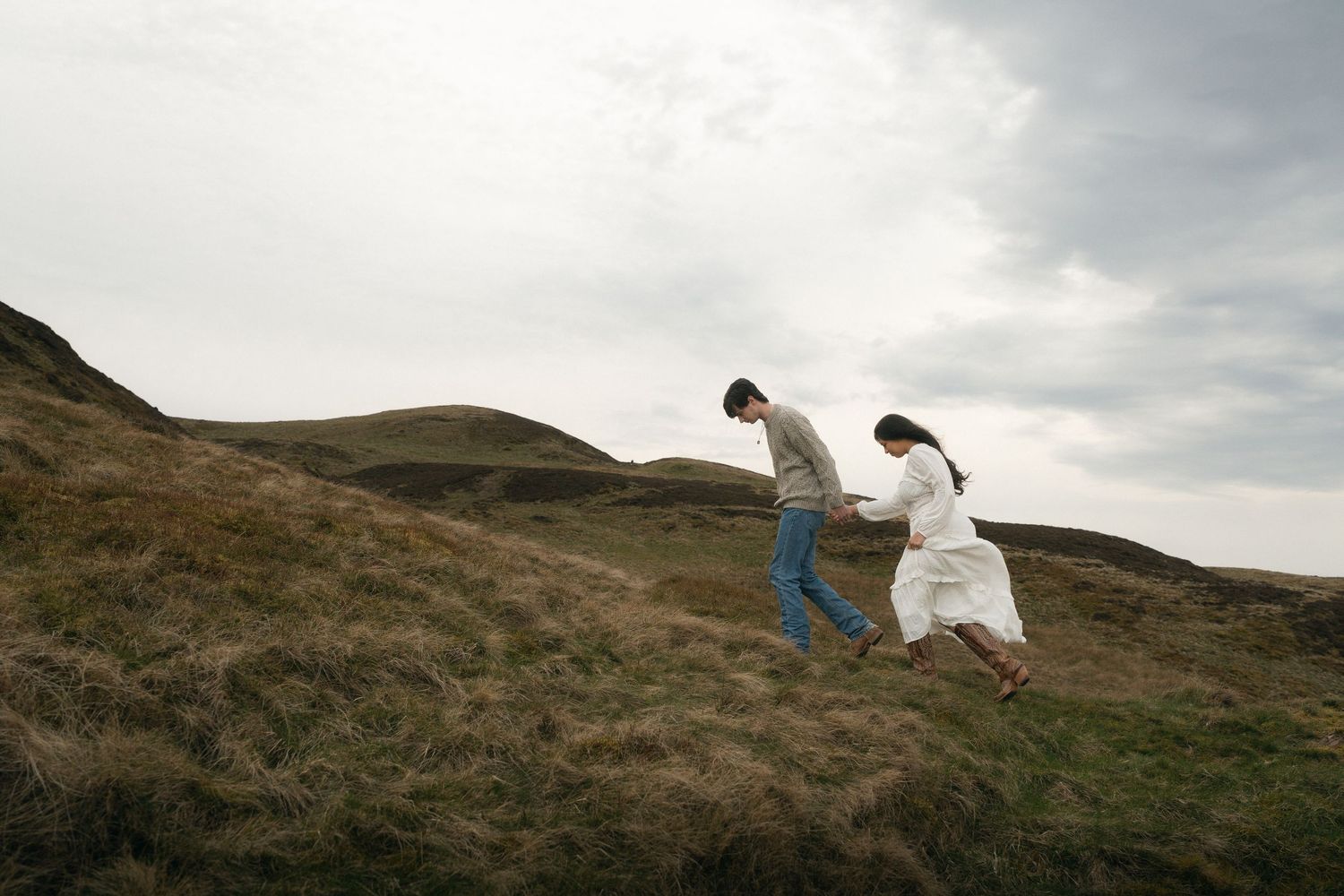 Couple holding hands walking up a grassy hillside on a cloudy day in a scenic landscape.