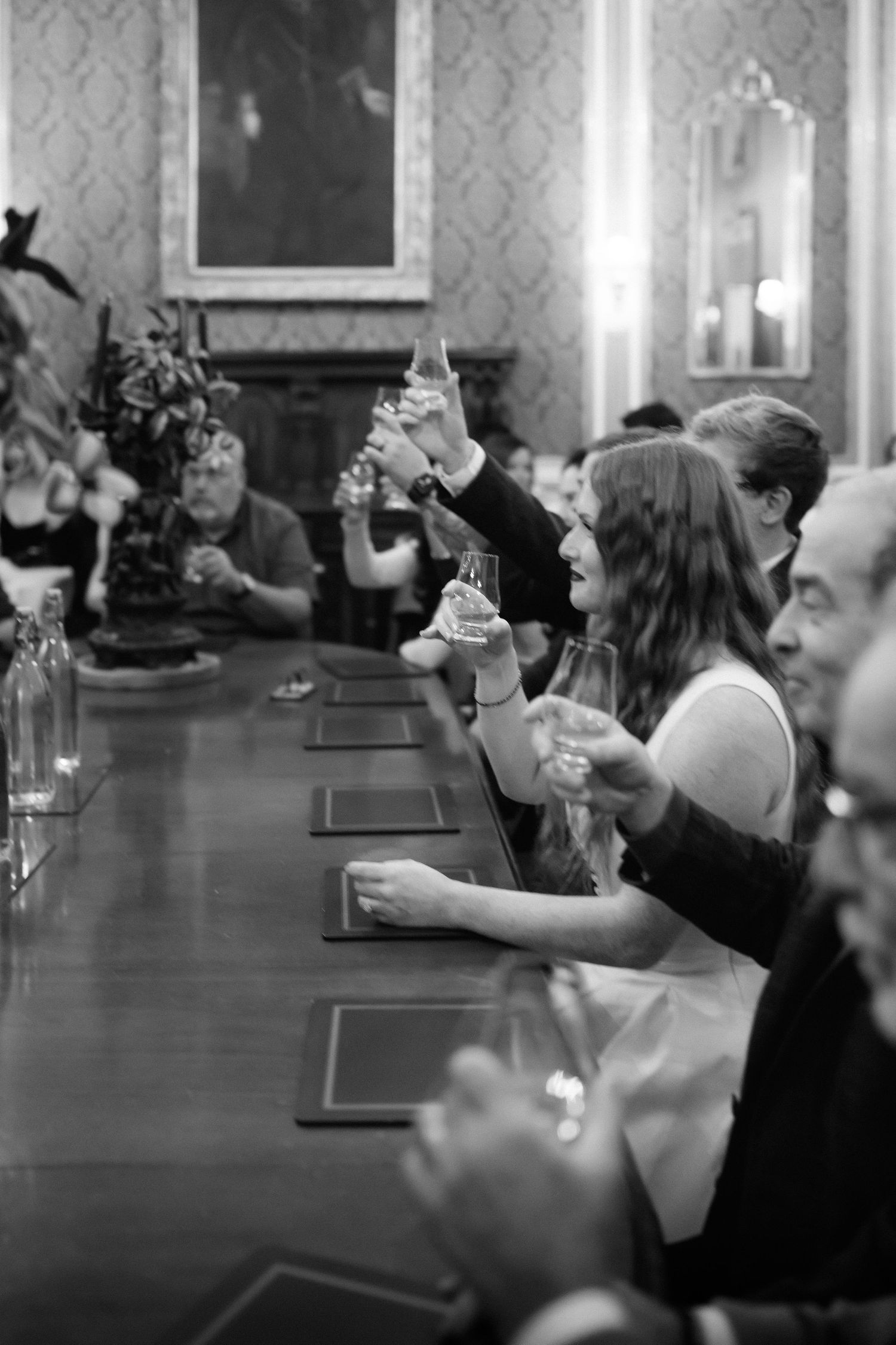 Guests raise their glasses in a toast during a formal dinner at an elegant dining room with patterned wallpaper.