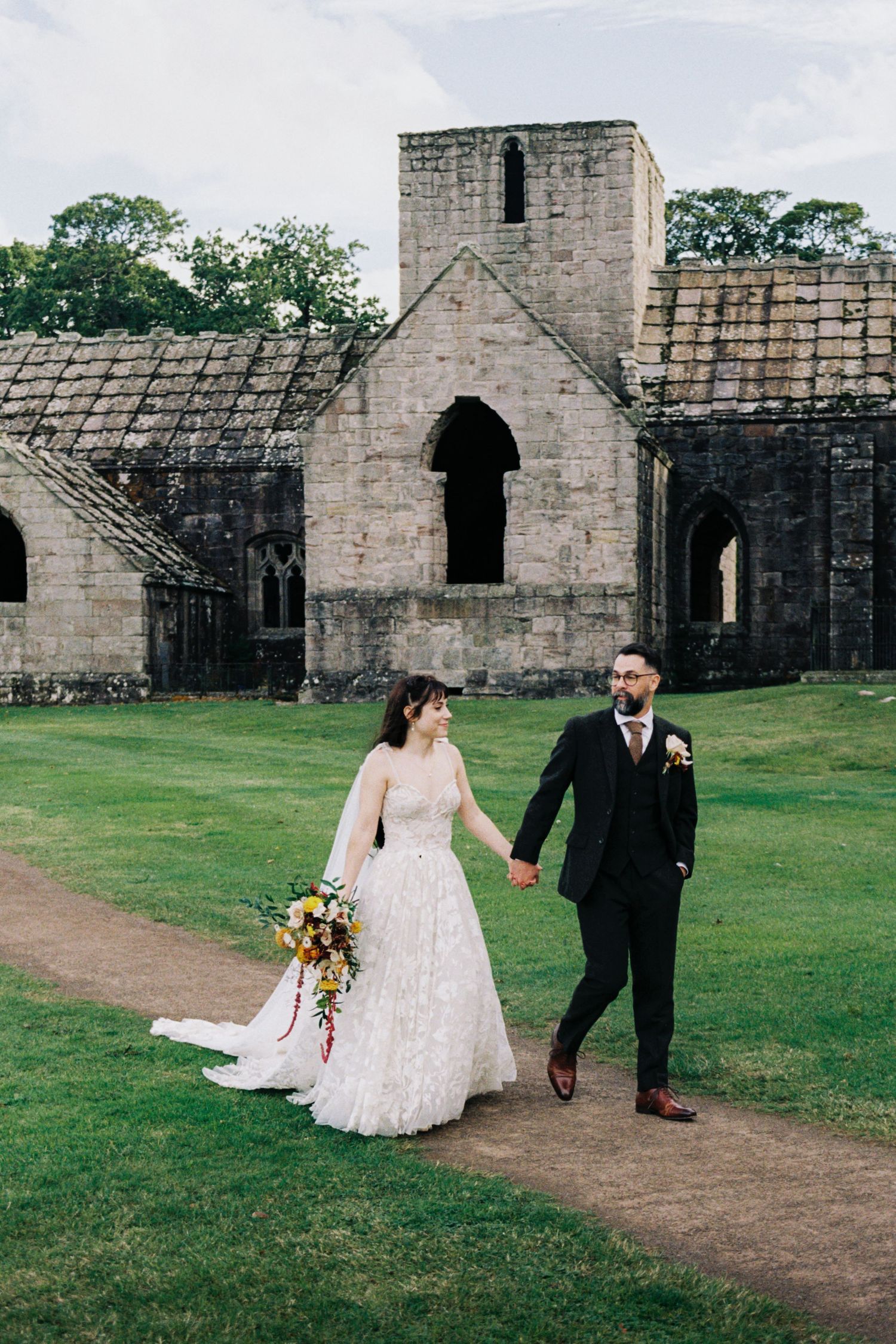 A couple walks together on a path near historic stone castle ruins during their wedding celebration.